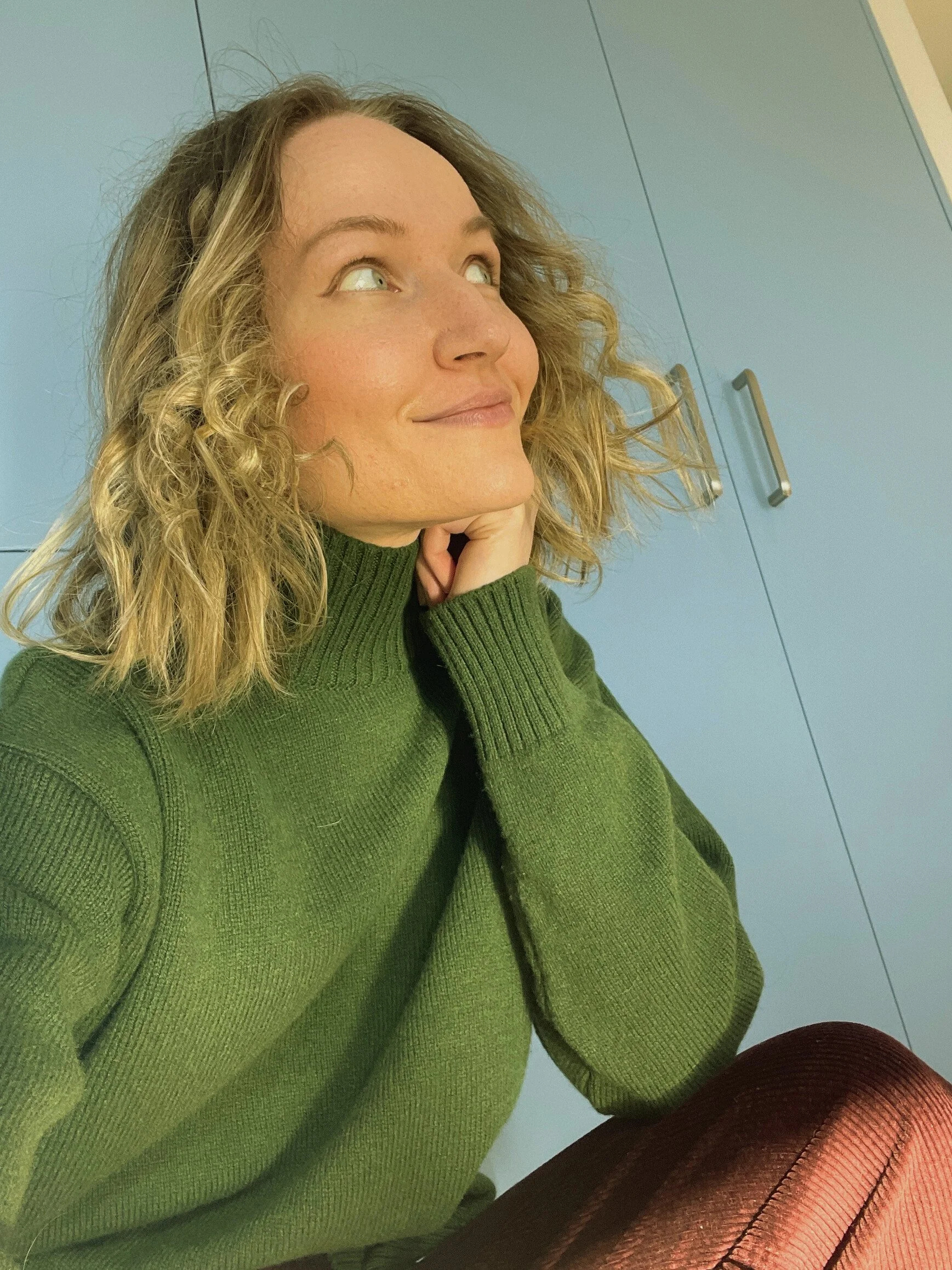 A woman with wavy blonde hair wearing a green turtleneck sweater, sitting in front of blue cabinets, looking to her left with a thoughtful expression.