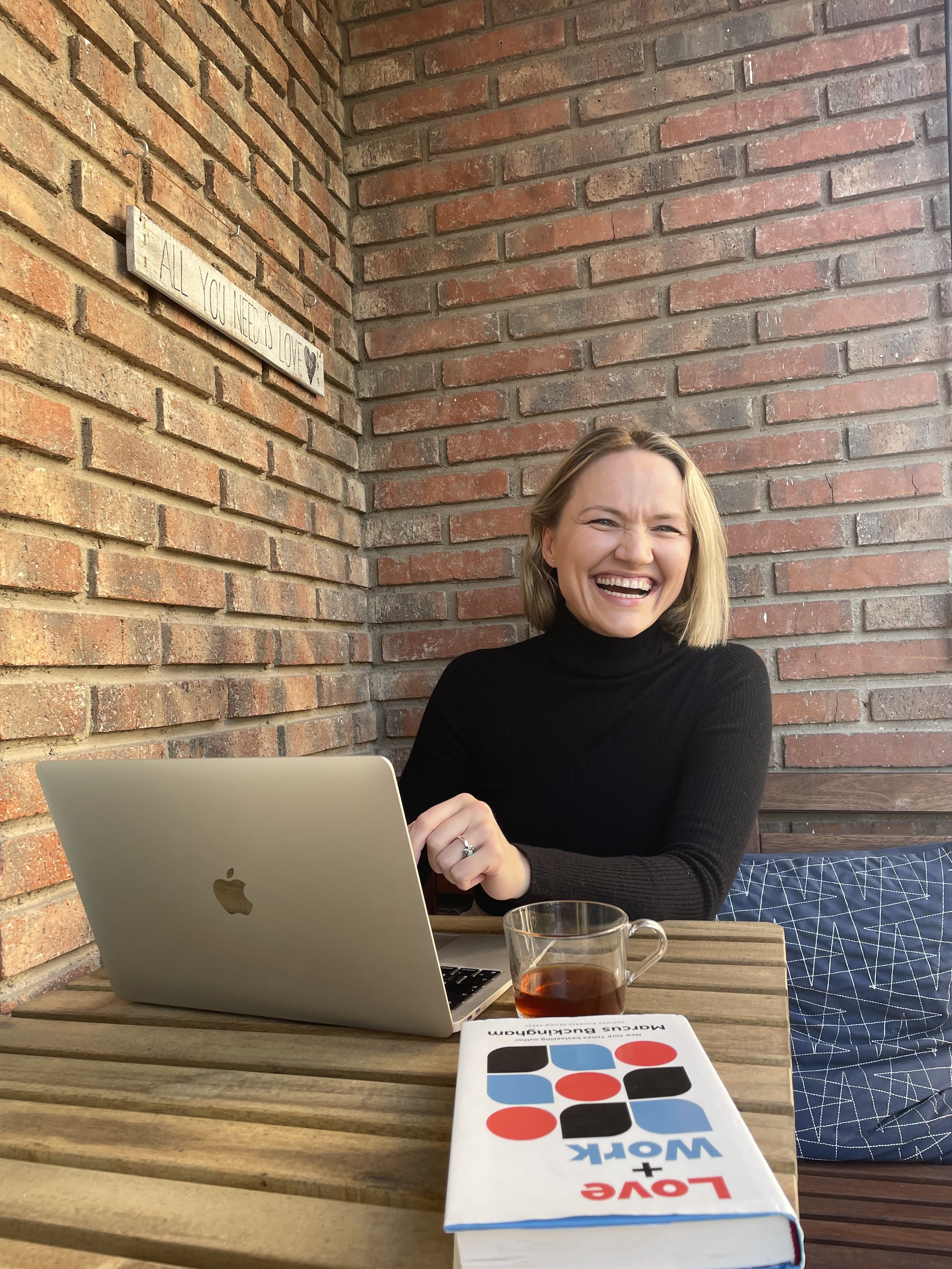 A woman in a black turtleneck sitting at a wooden table near a brick wall, smiling and looking to her right, with a MacBook laptop, a glass of tea, and a book titled "LOVE + WORK" on the table.