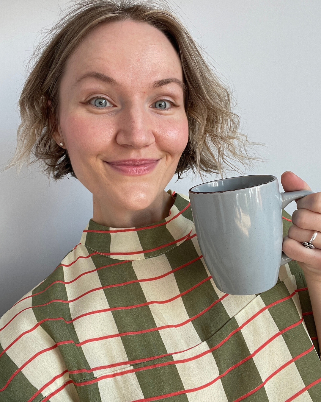 A woman with short, wavy blonde hair, blue eyes, and fair skin, smiling while holding a gray mug. She is wearing a green and beige checkered top with red stripes.