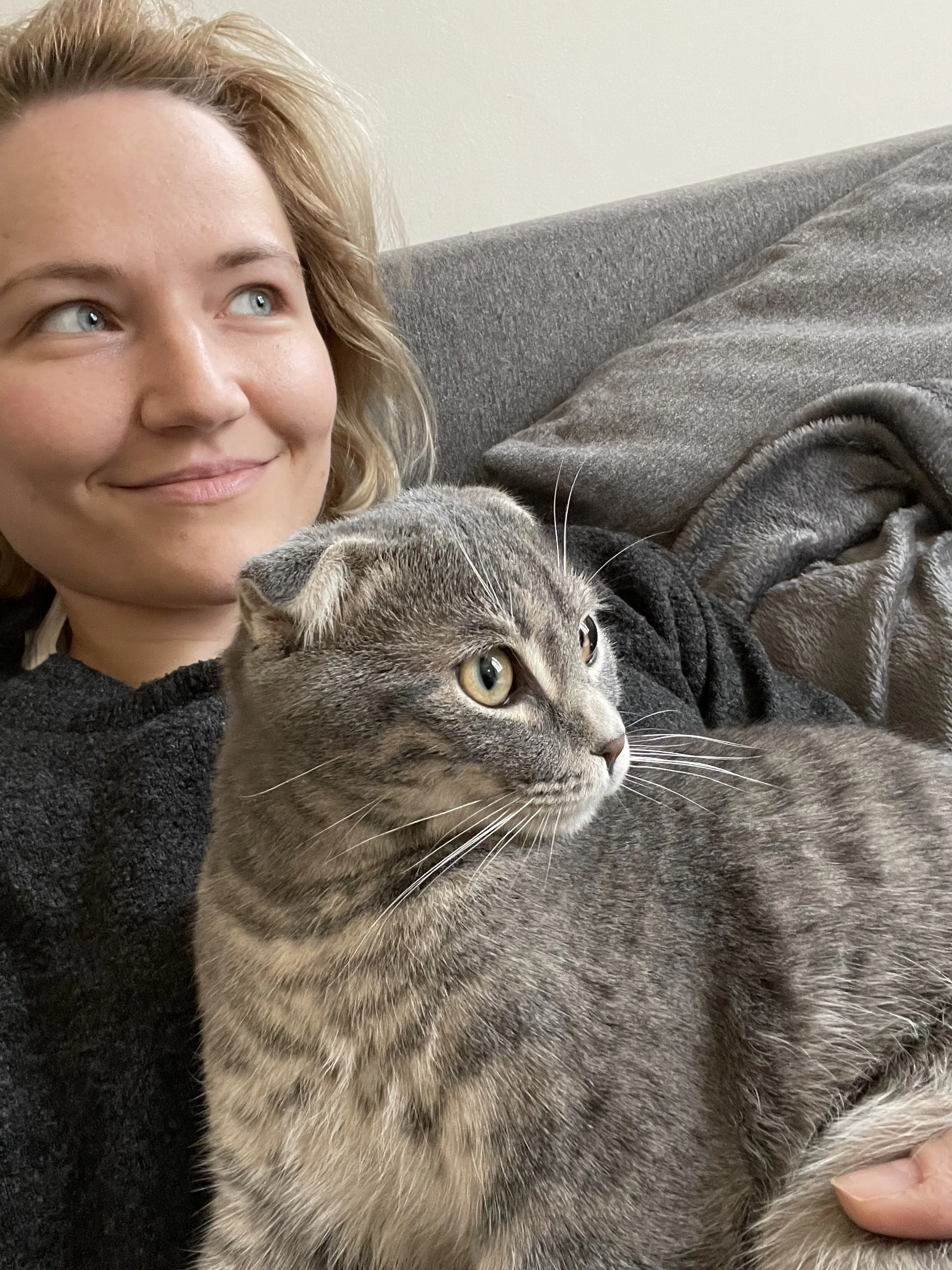 A woman with blonde hair resting on a gray couch, smiling, with a gray tabby cat lying on her chest, looking to the side.