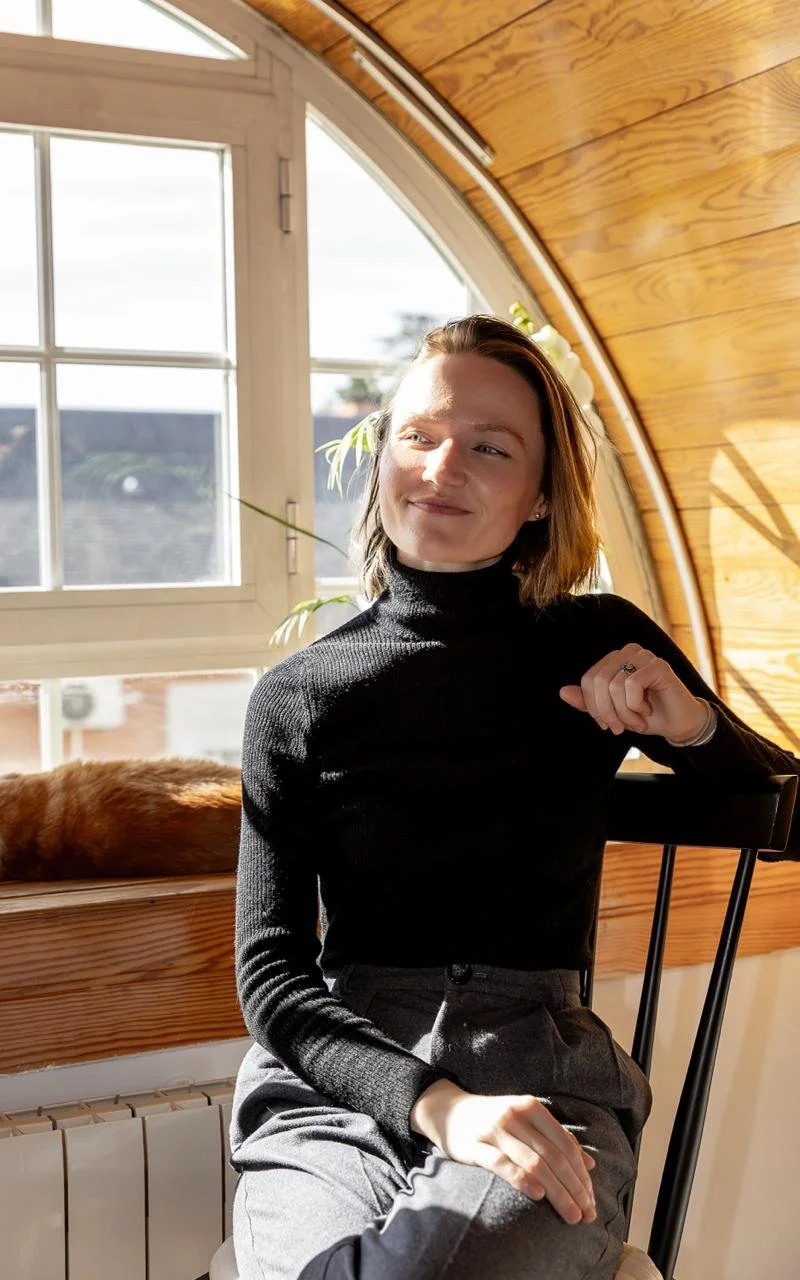 A woman with short brown hair sitting on a black chair inside a wooden-paneled room, with sunlight coming through a window behind her, and a cat resting on a ledge in front of the window.