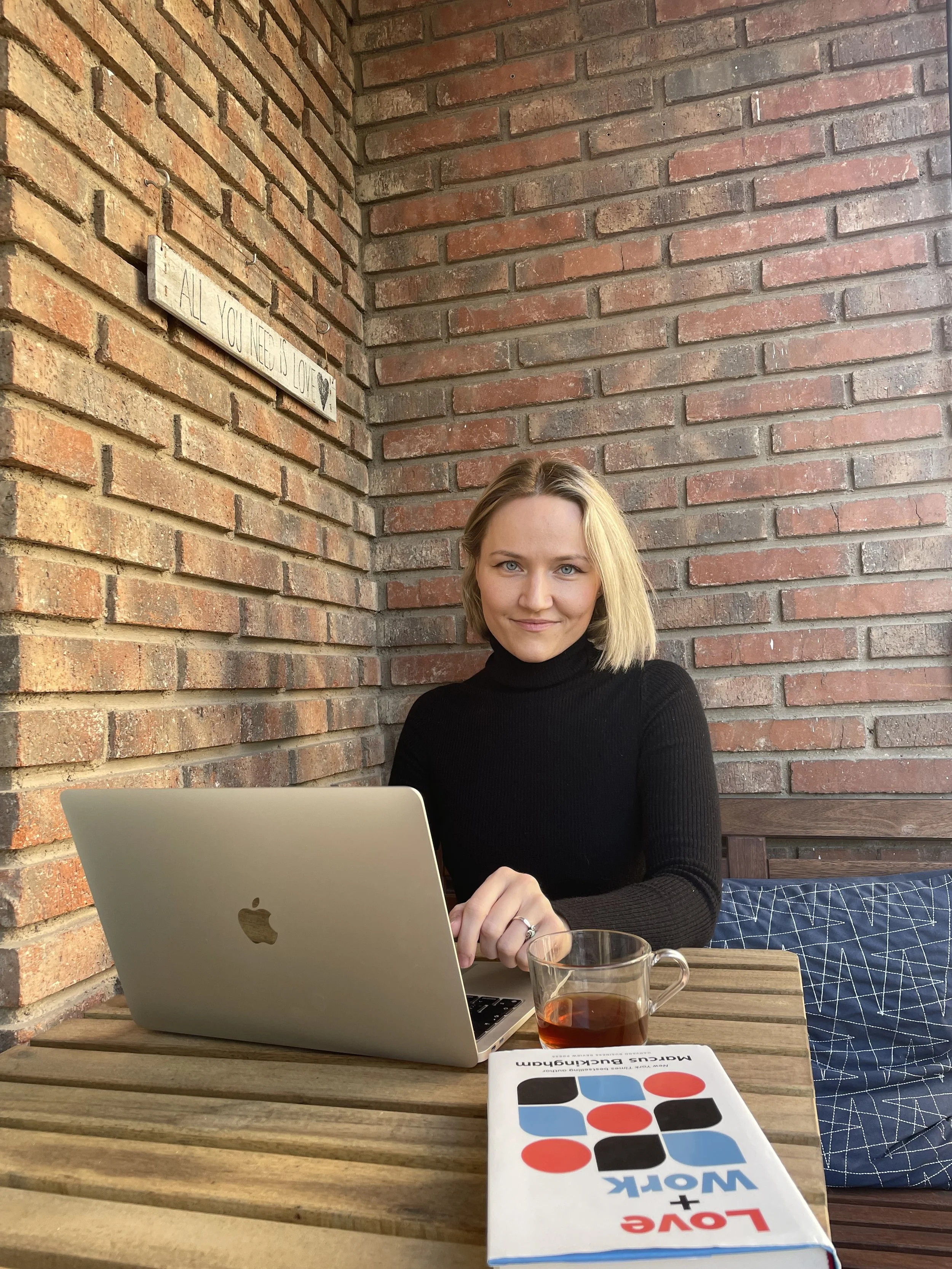 A woman with blonde hair and blue eyes sitting at a wooden table with a silver MacBook, a glass of tea, and a book titled 'Love + Work'. She is wearing a black turtleneck and smiling at the camera, with a brick wall background and a wooden sign that reads 'All you need is love' behind her.