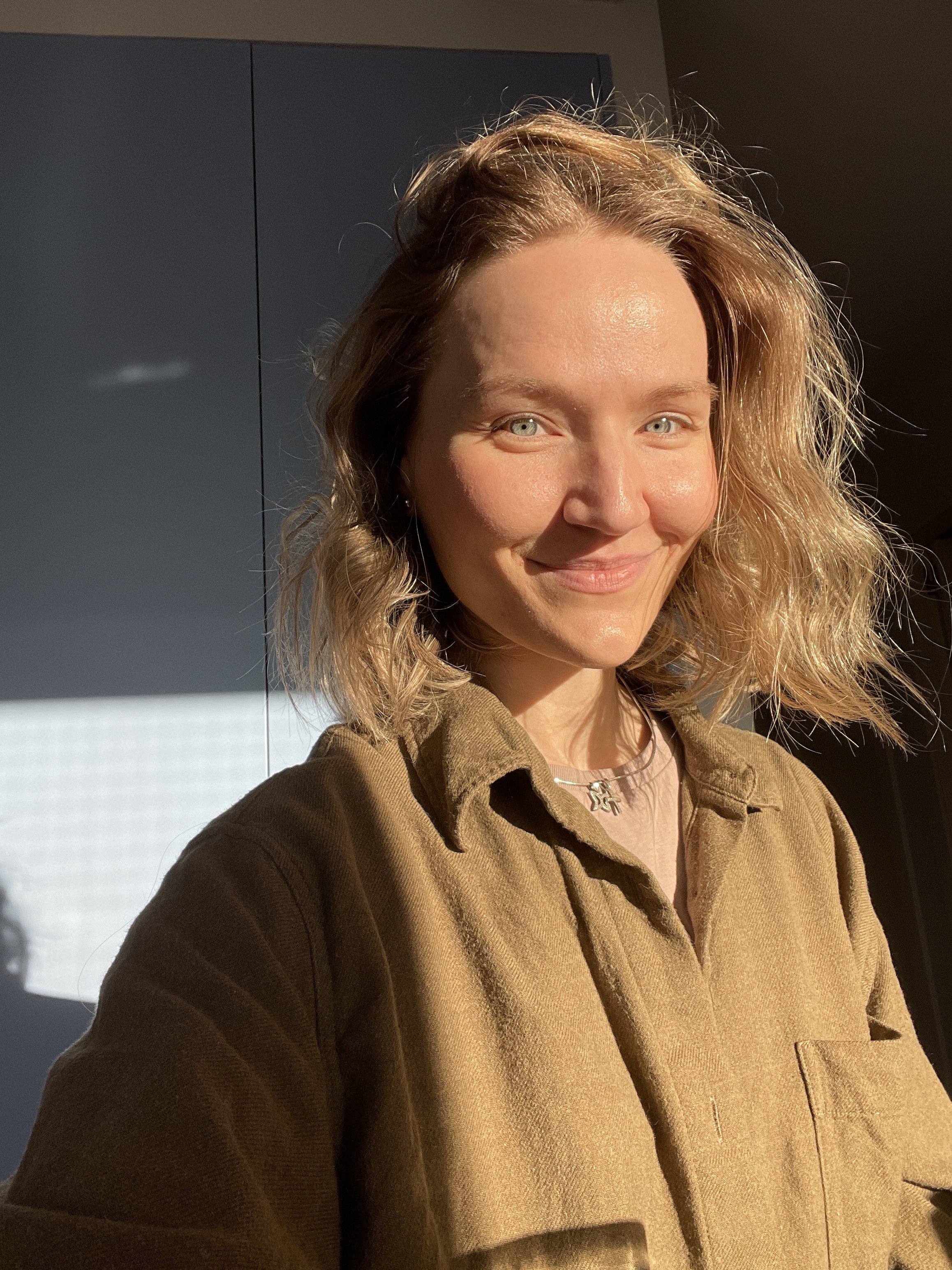 A woman with wavy blonde hair smiling at the camera with sunlight illuminating her face, wearing a brown shirt and a necklace, indoors.