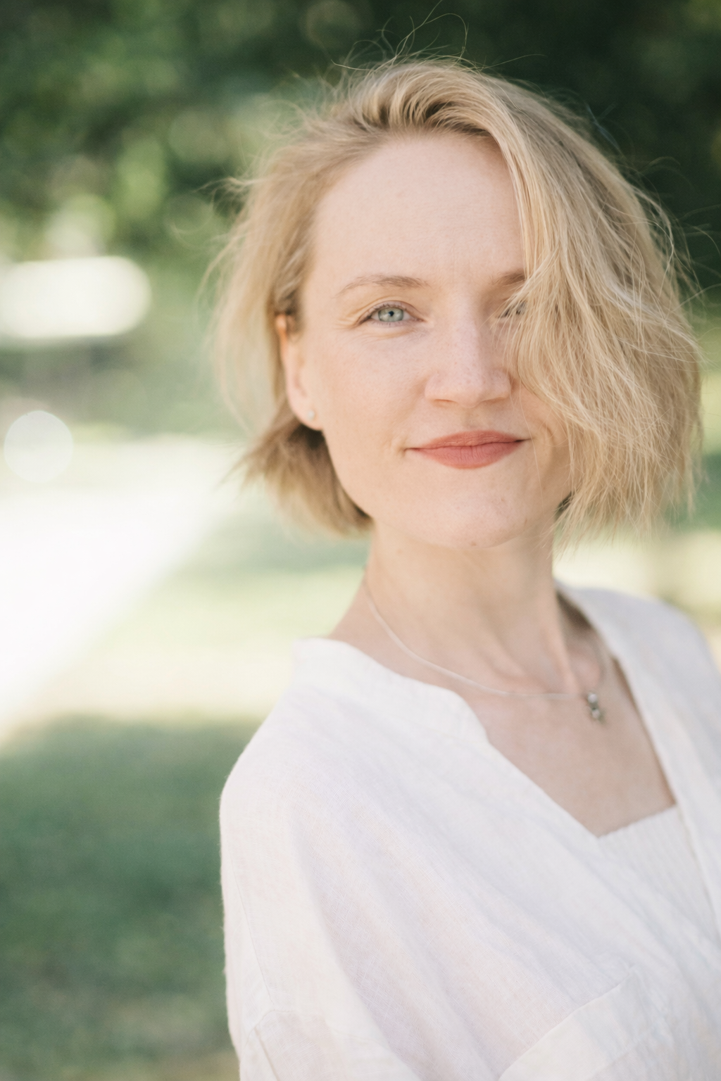 A woman with blonde, wavy hair and blue eyes smiling outdoors, wearing a white top and a delicate necklace.