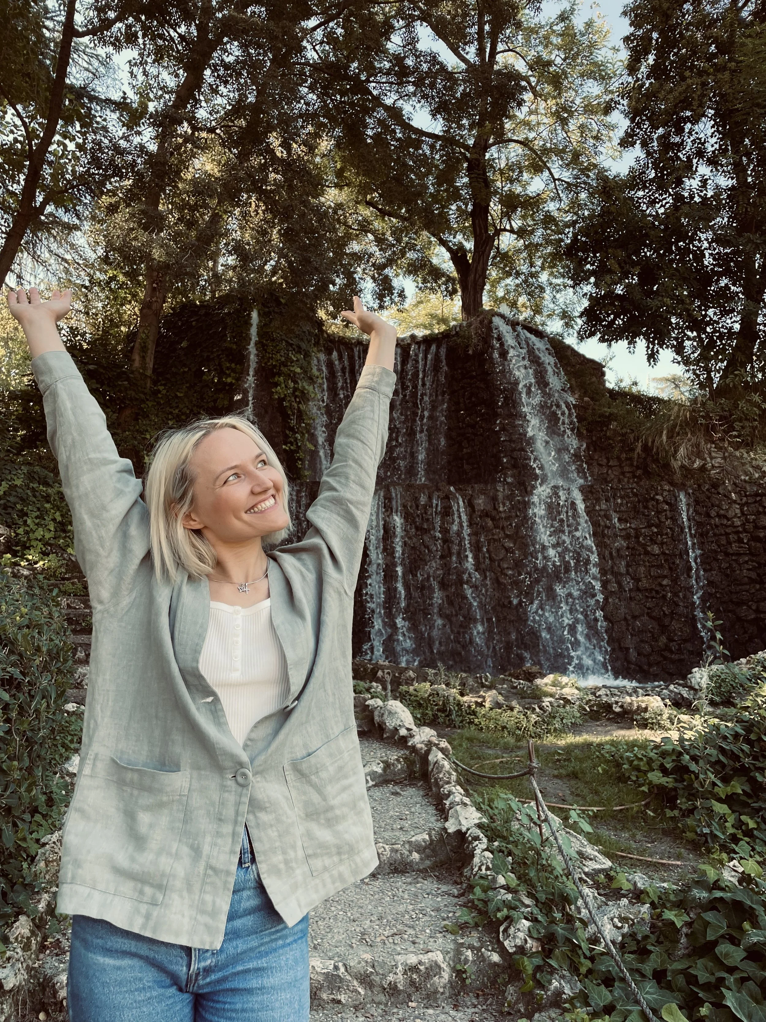 A woman with blonde hair, smiling and raising her arms in front of a waterfall surrounded by trees and greenery.