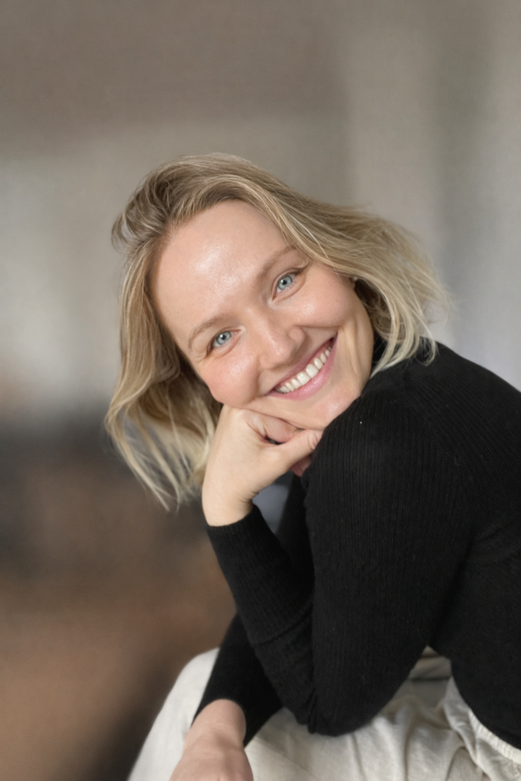 A woman with blonde hair and blue eyes smiling, resting her chin on her hand, wearing a black long sleeve shirt, against a blurred background.