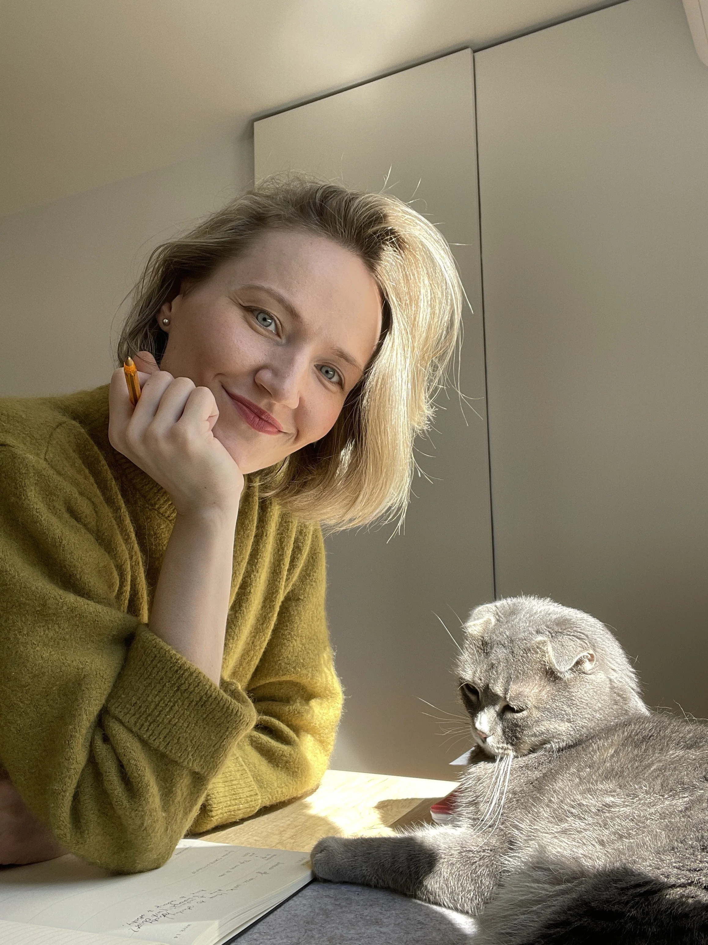 A woman with blonde hair and light blue eyes wearing a green sweater, resting her chin on her hand and smiling at the camera, sitting next to a gray cat with folded ears and closed eyes, laying on a desk with a notebook nearby.