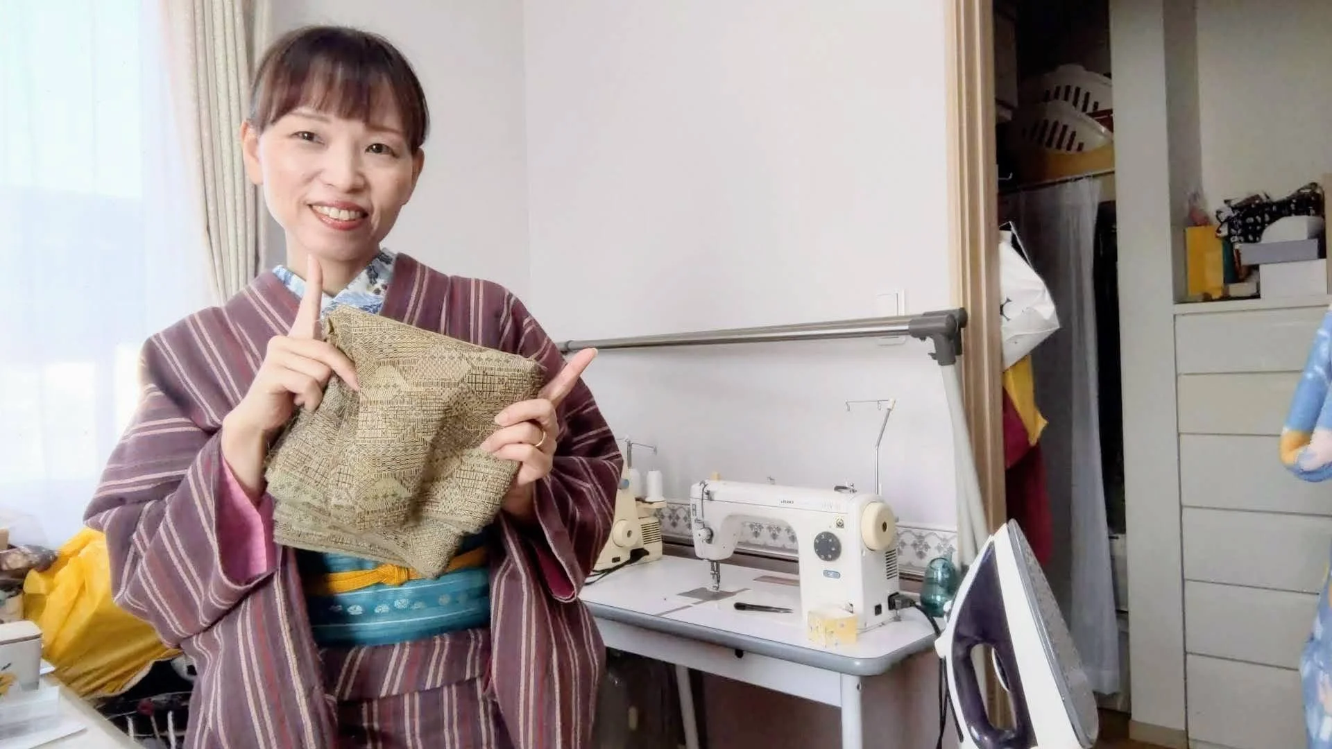 縞模様の着物を着た女性が笑顔で手に布を持ち、指を立てている。背景には縫い機や衣服、収納棚が見える室内の風景。