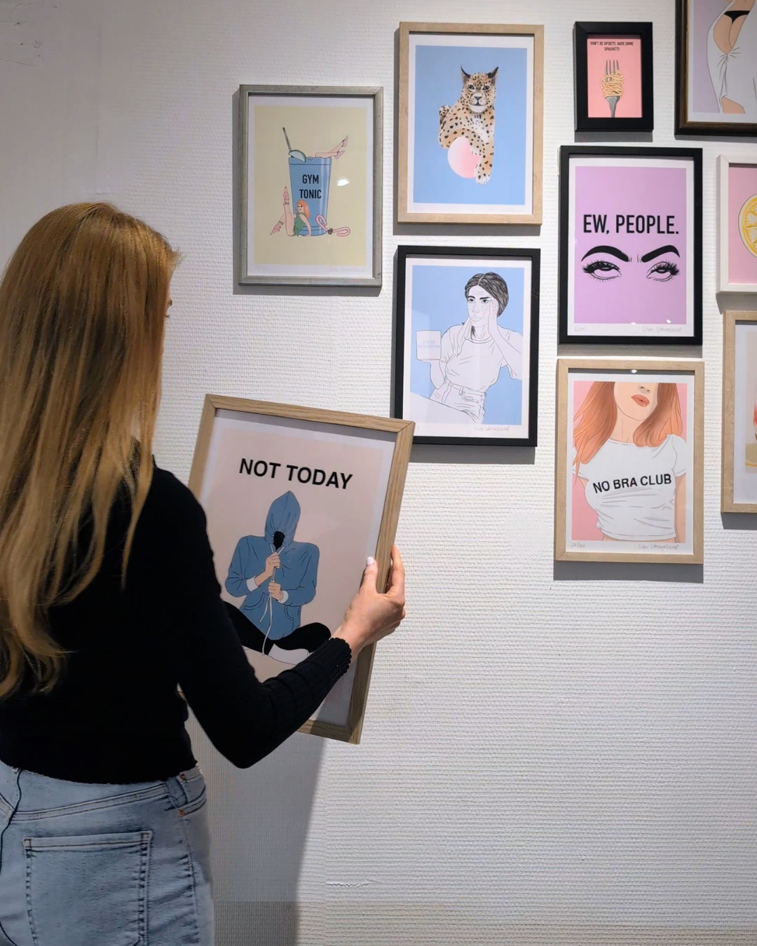 A person with long blond hair holding a framed picture that says "NOT TODAY" with an illustration of a person with a hoodie and a microphone. The person is standing in front of a white wall with various framed colorful illustrations and quotes, including a cheetah, a woman with red hair, and stylized eye drawings.