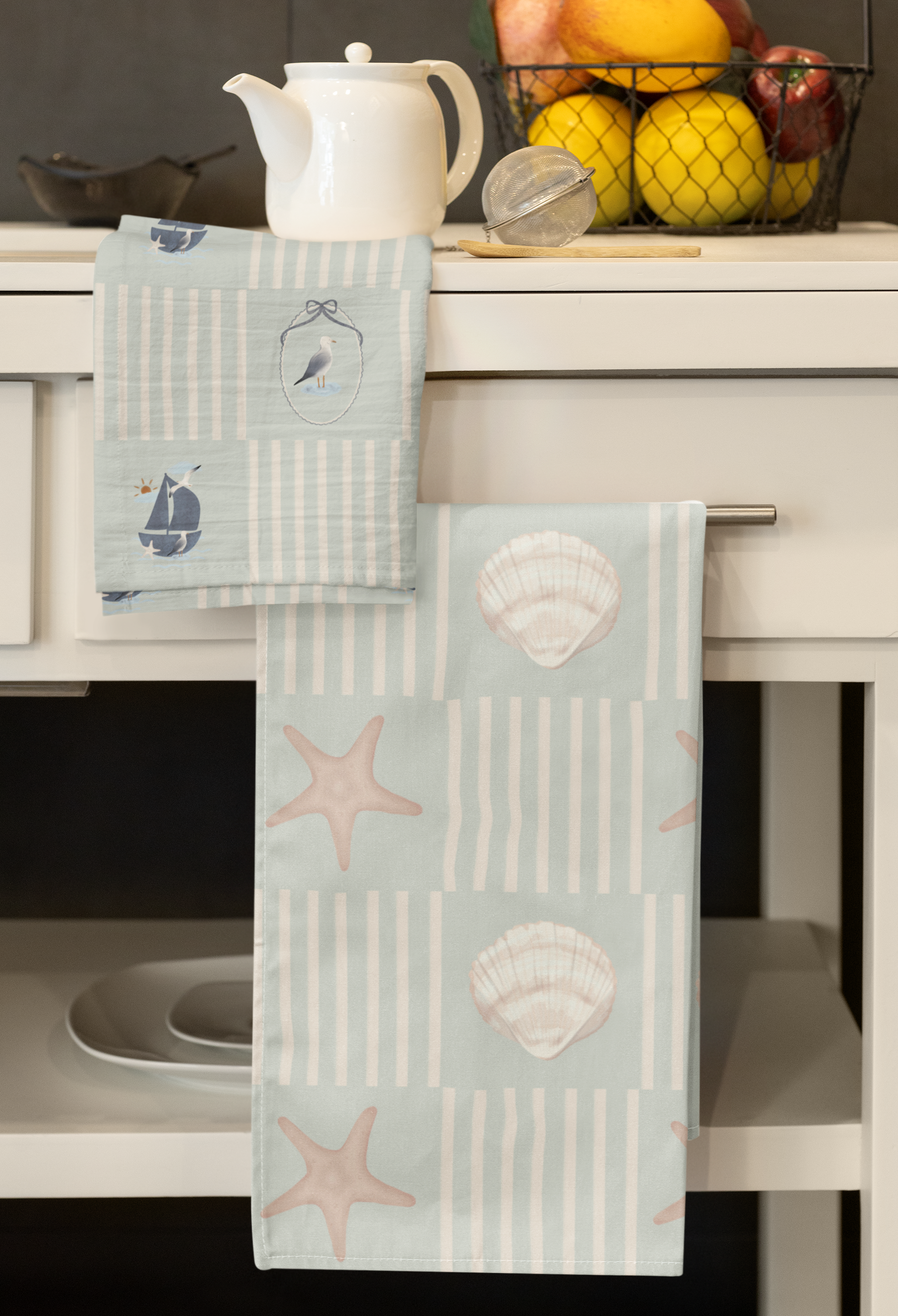 A white kitchen counter with a white ceramic teapot, a mesh fruit basket filled with apples and citrus fruits, a metal tea infuser, and striped kitchen towels with seashell and starfish patterns hanging over the counter edge.