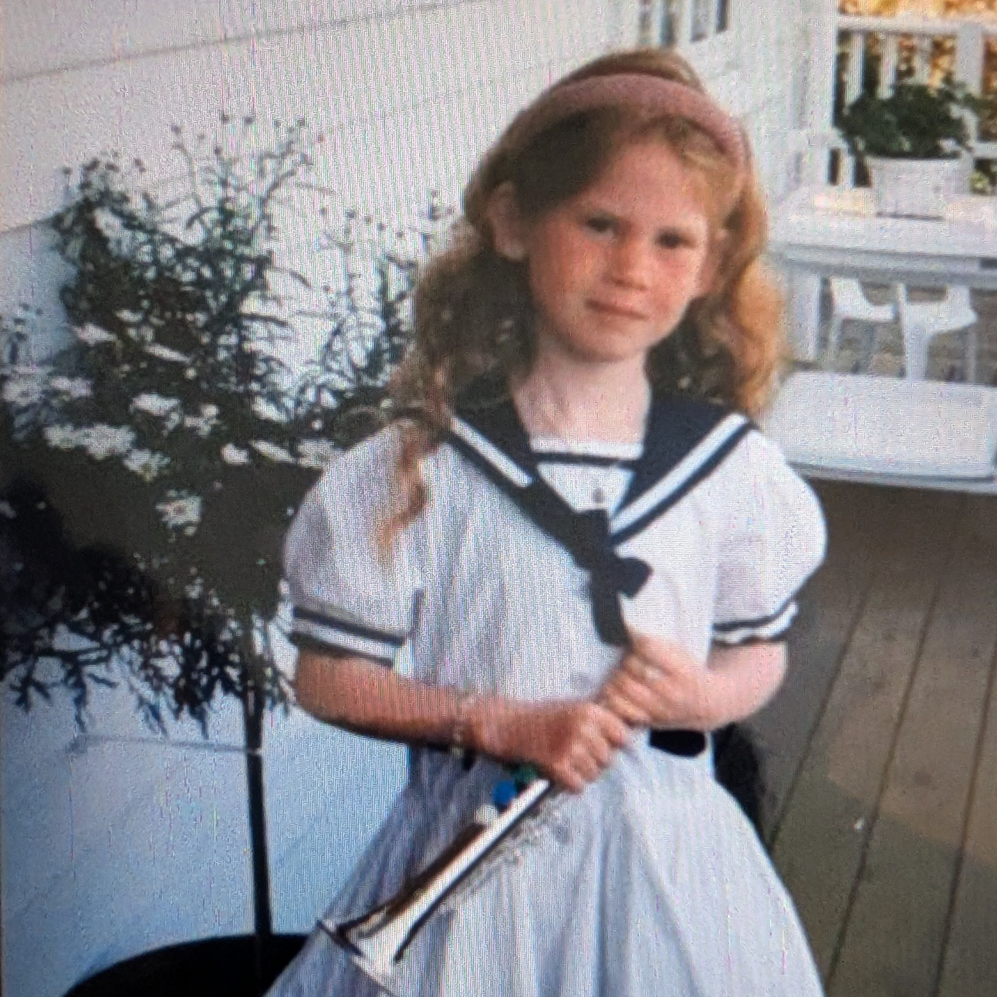 A young girl with reddish curly hair, wearing a white dress with navy accents, holding a white umbrella, standing in front of a potted plant and a porch with white railings.