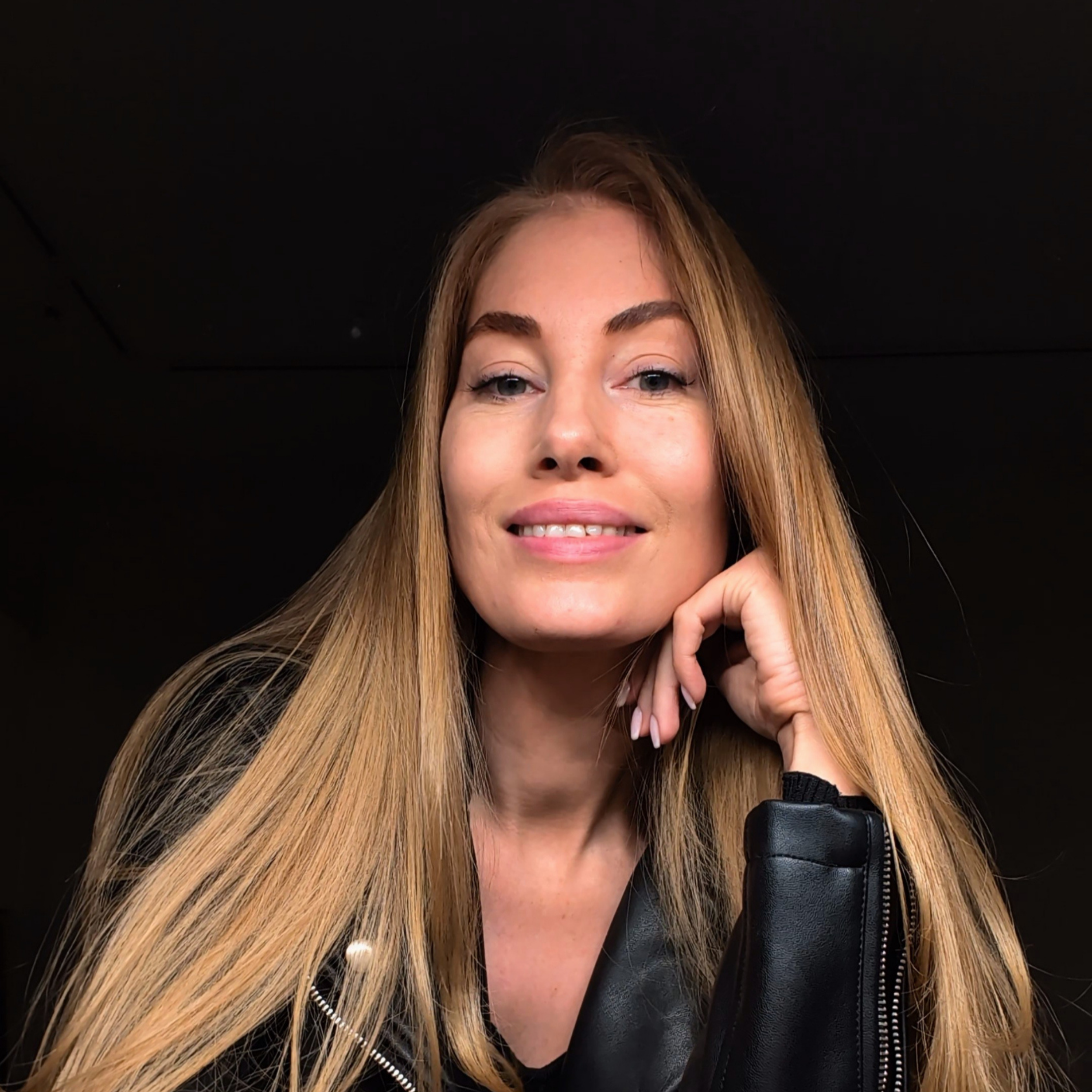 A woman with long blonde hair, light skin, and a black leather jacket smiling and resting her chin on her hand against a dark background.