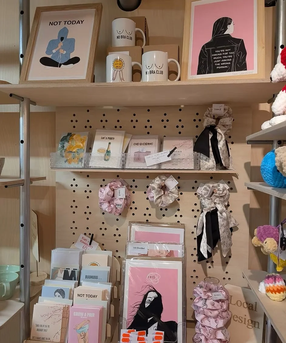 Decorative store display with framed prints, mugs, scrunchies, greeting cards, and small yarn accessories arranged on shelves and a pegboard.