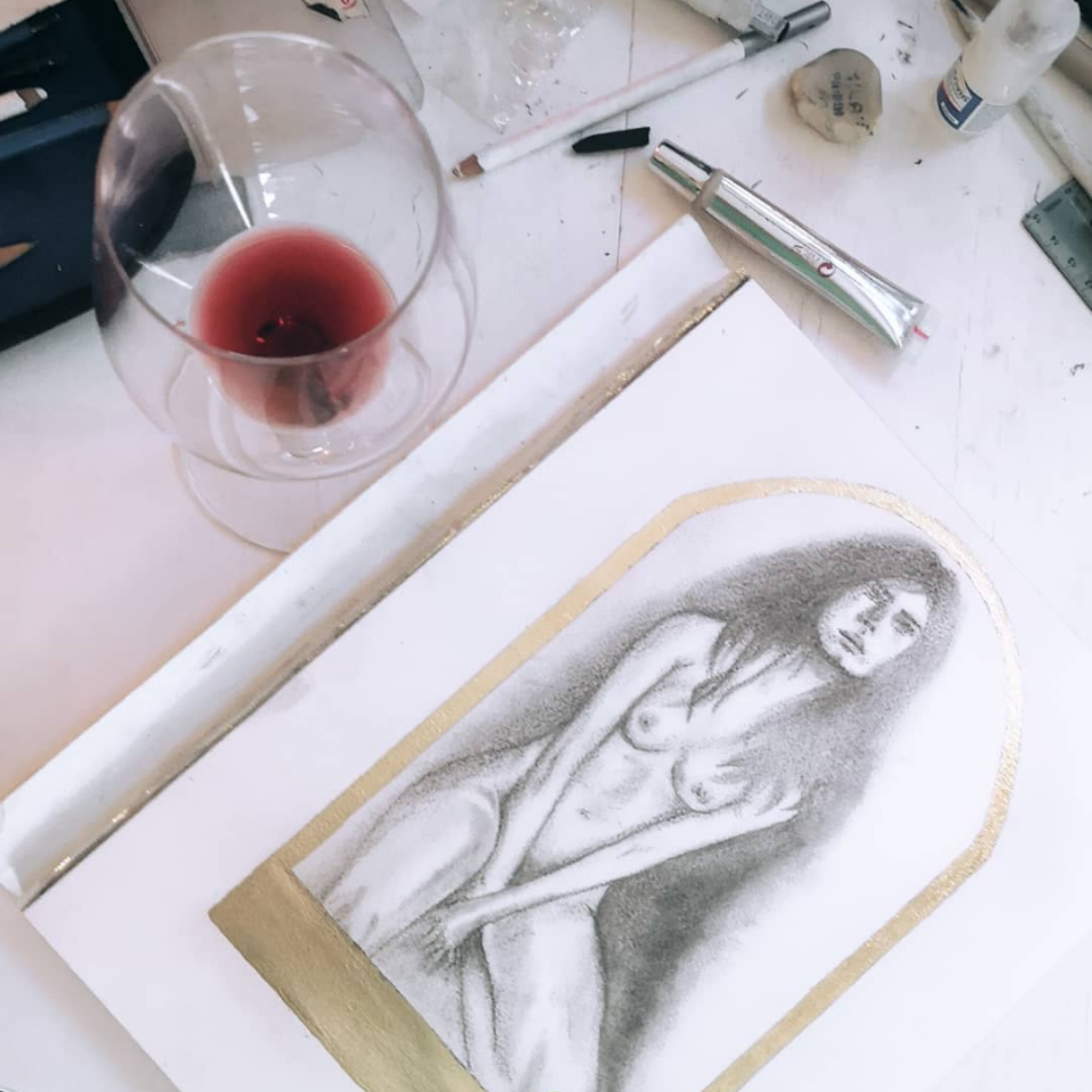 A realistic pencil drawing of a nude woman on white paper, bordered with gold, placed on a cluttered white table with a glass of red wine, art supplies, and tools.