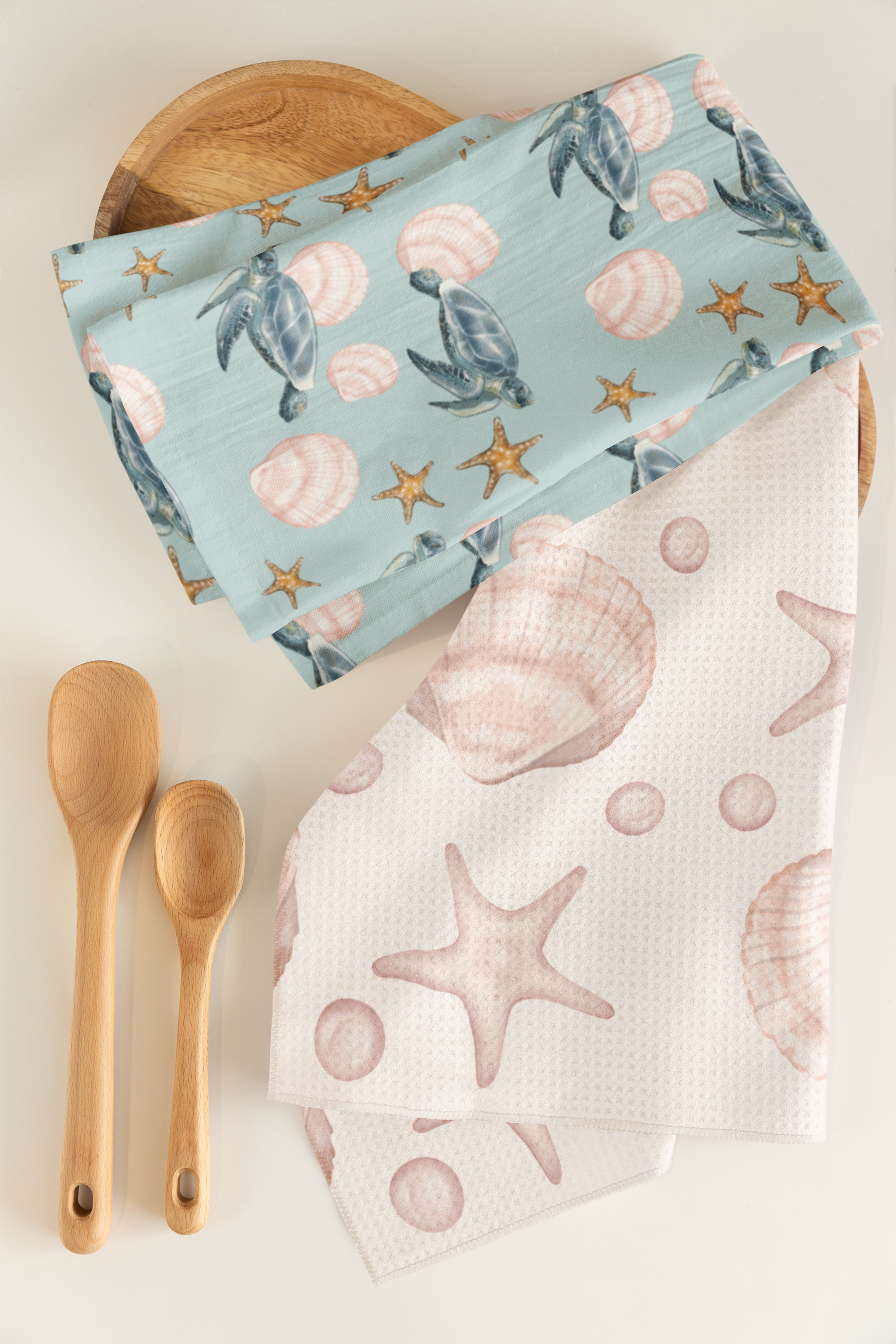 Beach-themed fabric napkins with shells, starfish, and sea turtle designs, two wooden spoons, and a round wooden tray on a white surface.