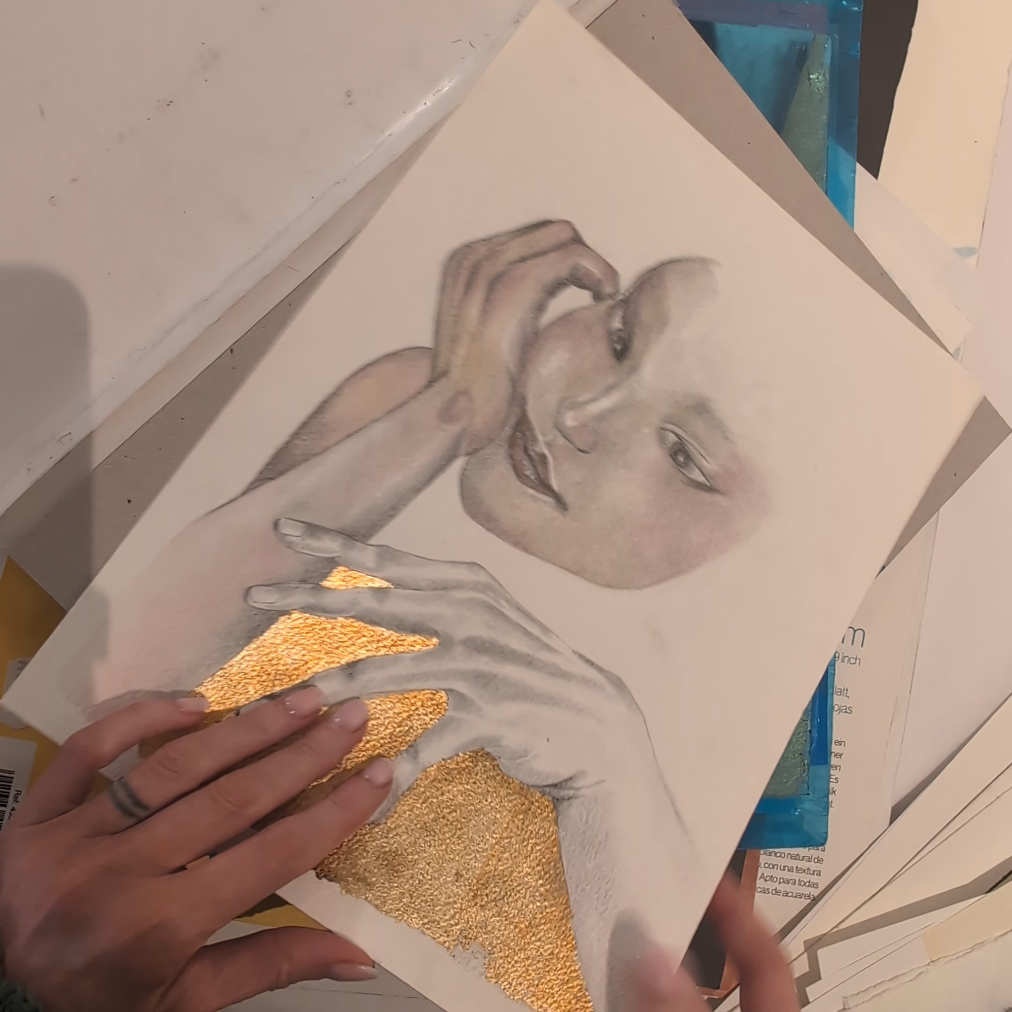 A hand holding a drawing of a woman’s face and hands, with gold-colored accents on the hands and clothing. The woman has expressive eyes and a calm expression.