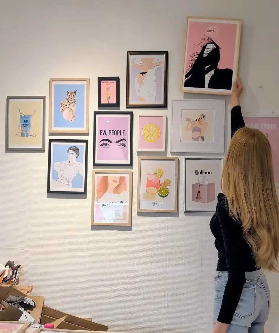 A woman with long blonde hair wearing a black top and light blue jeans is hanging up a framed art print on a wall decorated with various other framed illustrations and posters. The wall features colorful, stylish artwork, including portraits, text, and graphic designs.