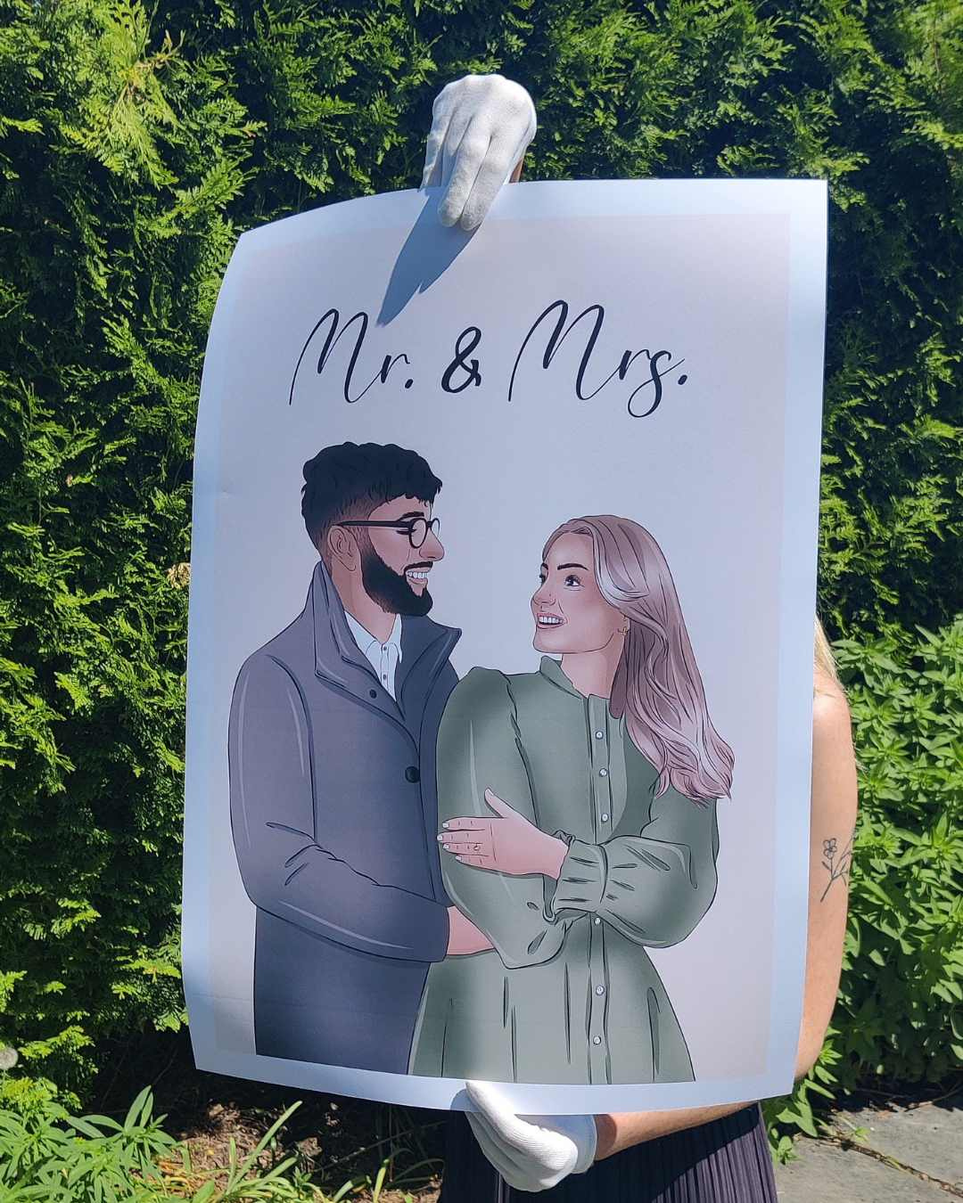 Illustration of a couple, a man with glasses and a woman with long blonde hair, smiling at each other, with the text 'Mr. & Mrs.' above them, held up by a person wearing white gloves in front of green bushes.