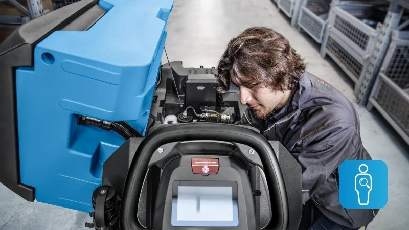 Technician inspecting or repairing a piece of industrial machinery inside a warehouse or factory.