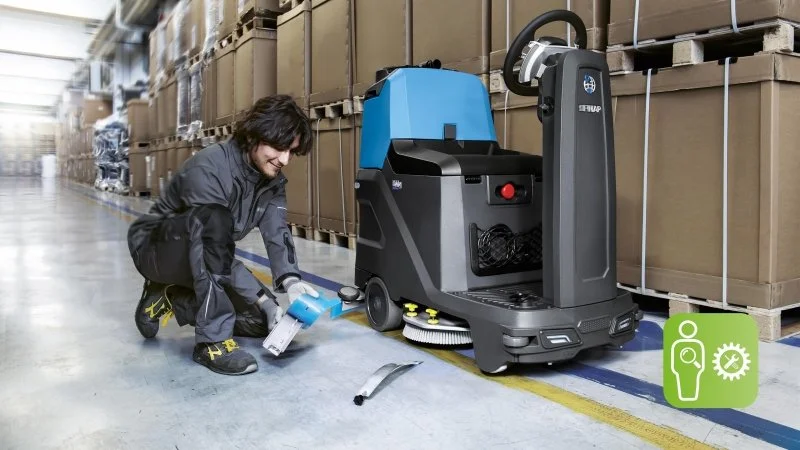 A man operating a floor cleaning or sweeping machine in a warehouse, with shelves filled with boxes in the background.