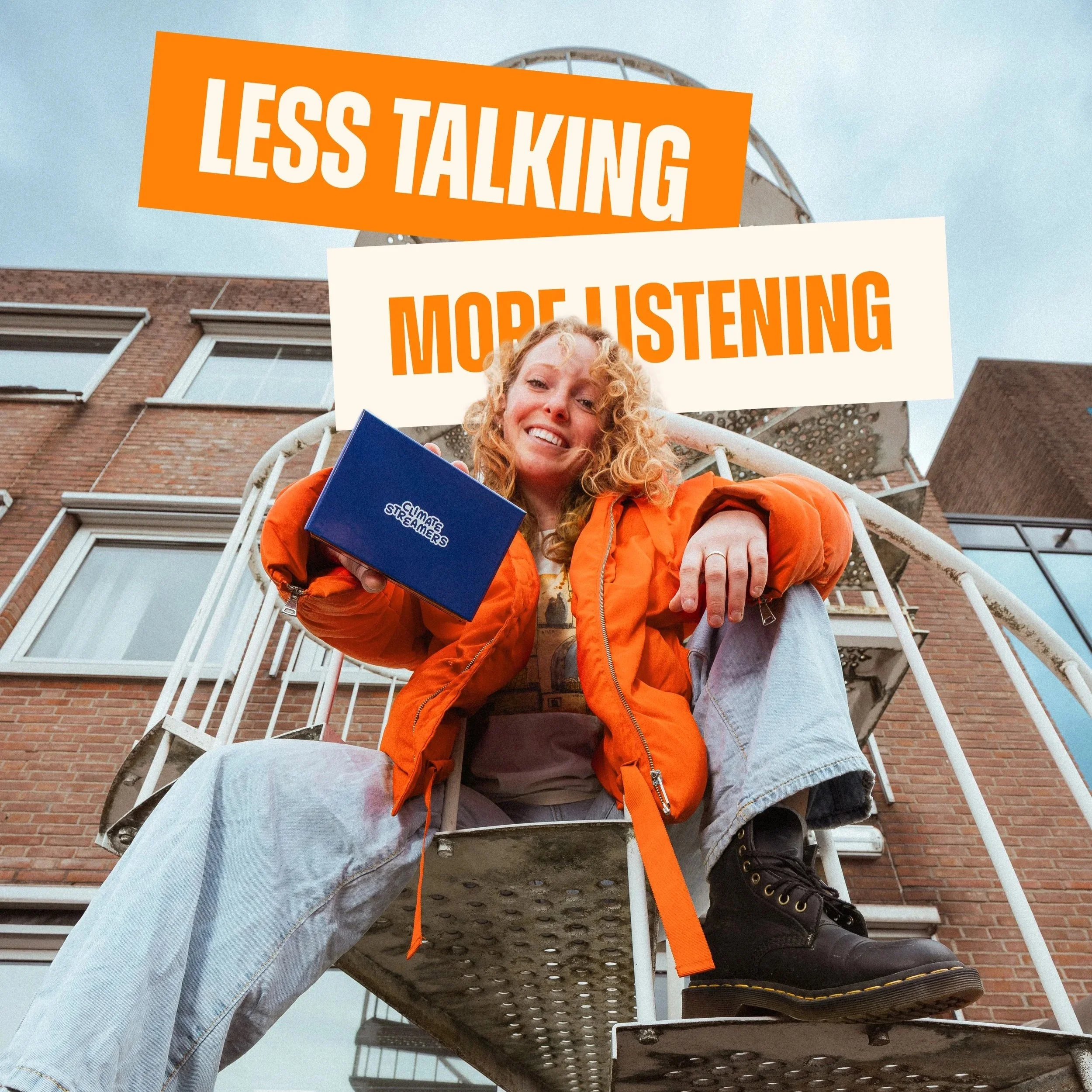 A woman with curly hair wearing an orange jacket and gray pants sitting on an outdoor metal staircase holding a blue booklet titled 'Climate Streamers,' with signs that read 'Less Talking' and 'More Listening', against a background of a brick buildin