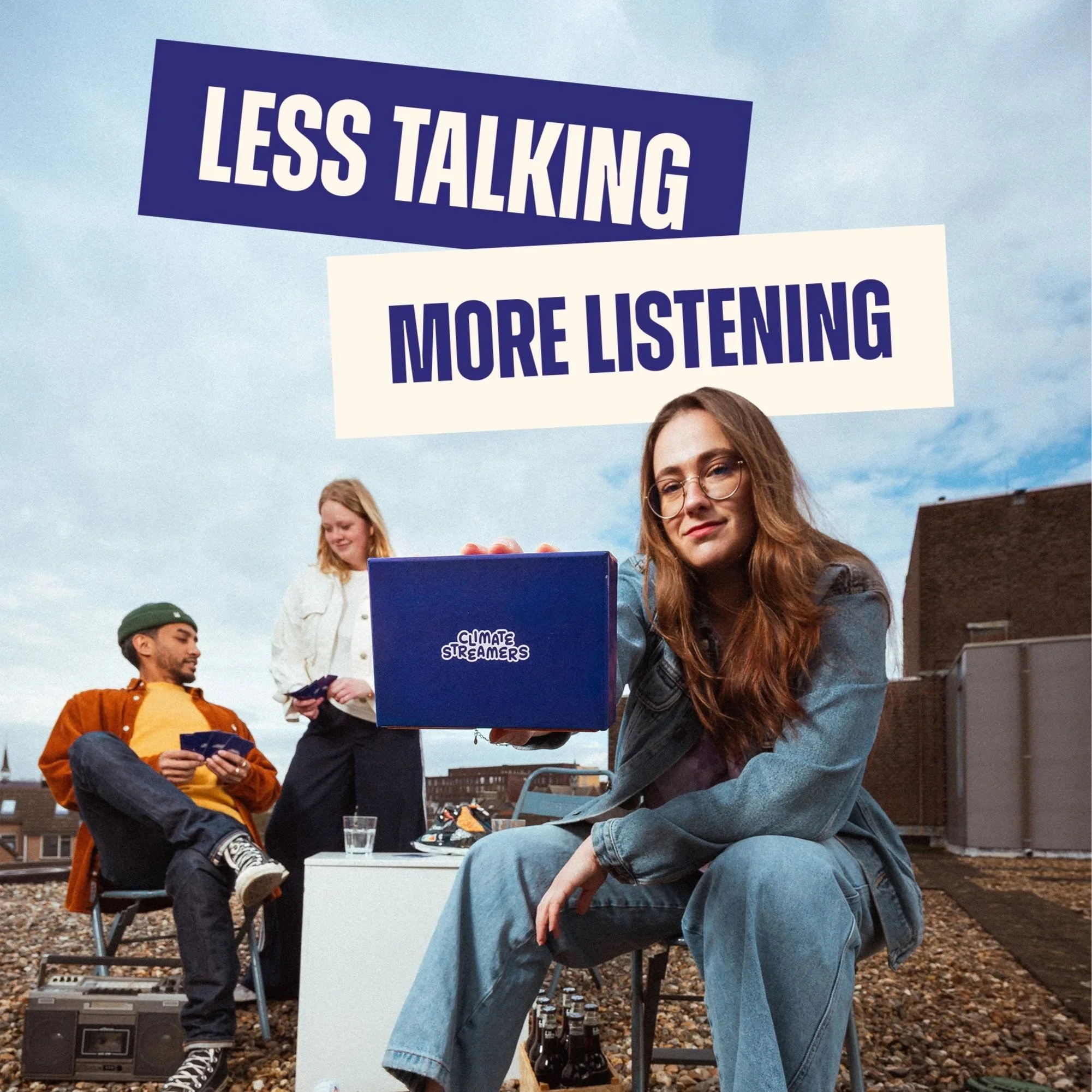Group of young adults outdoors at a social gathering, with one woman holding a large blue box labeled 'Climate Streamers' and a sign that reads 'Less Talking More Listening', suggesting an environment-focused event.