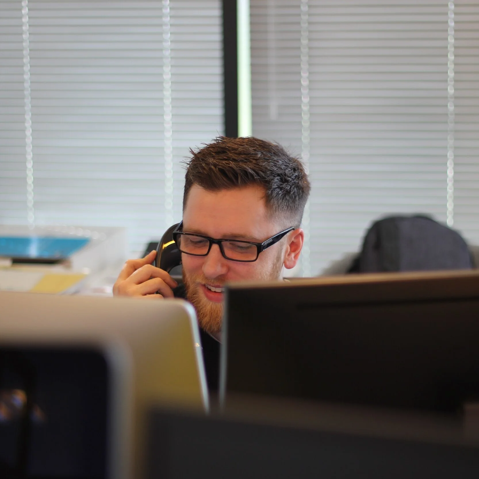 A man with glasses and a beard, talking on the phone in an office setting, with windows and blinds in the background.