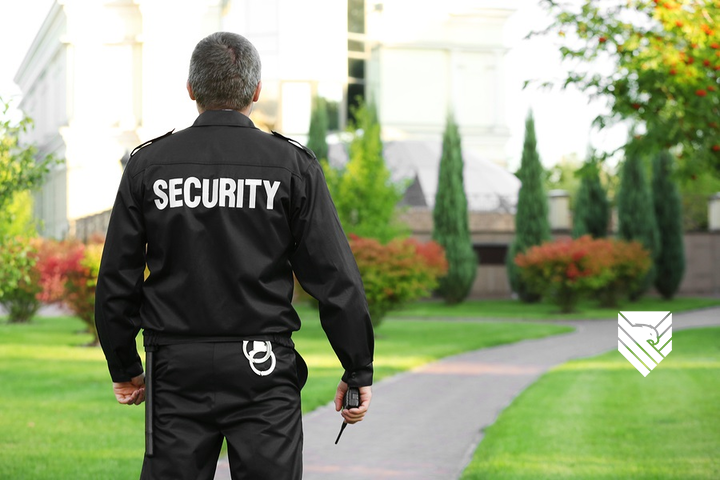 Security guard in black uniform holding a walkie-talkie, walking on a sidewalk in a residential area with trees and houses in the background.