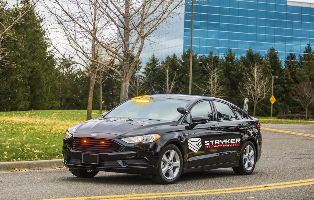 A black Ford sedan with a security company logo on the side, parked on a street near a park with trees and a modern glass building in the background.