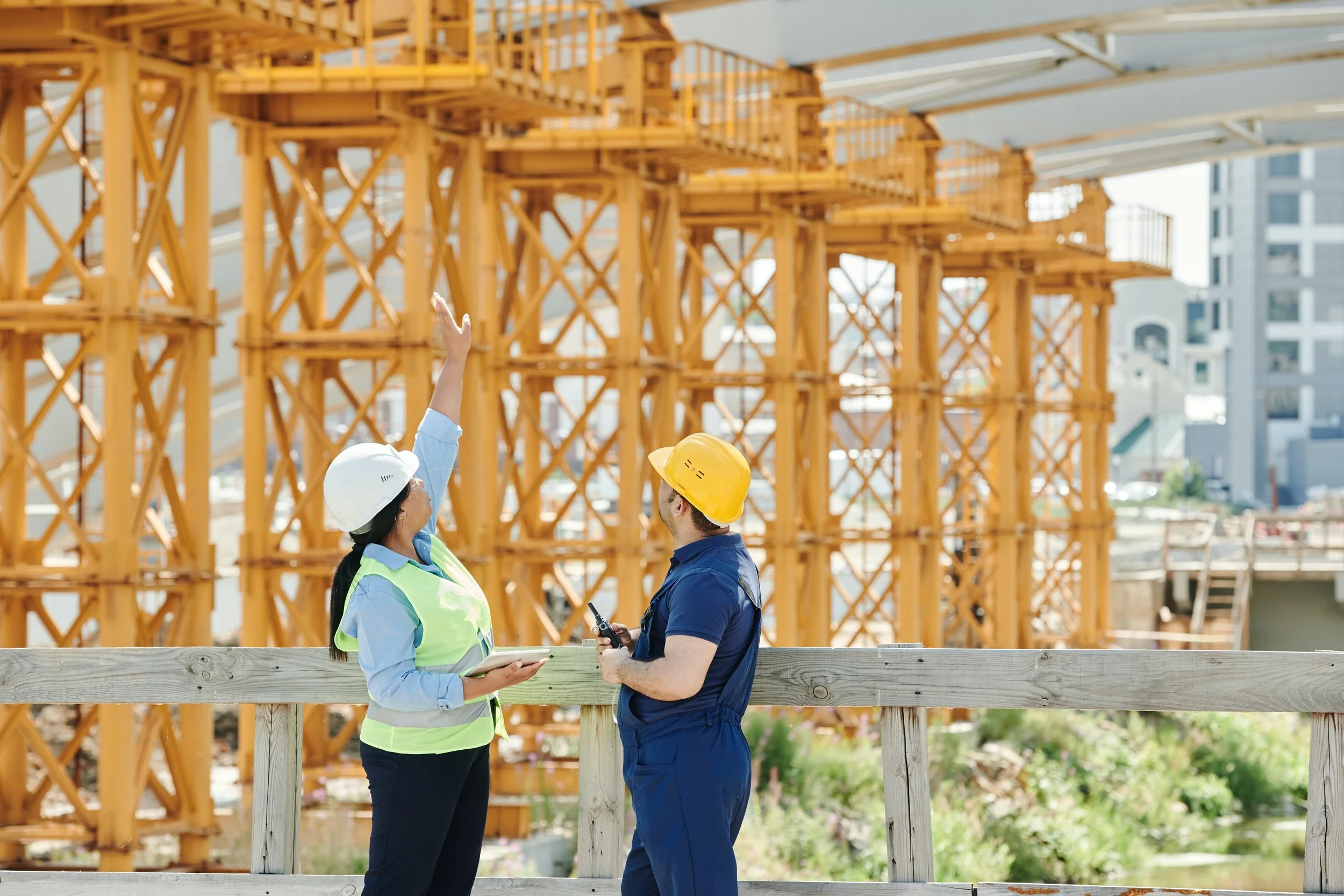 Two construction workers, a woman and a man, discussing at a construction site in front of a large orange crane structure. The woman is pointing upward while holding a tablet, and the man is holding a walkie-talkie.