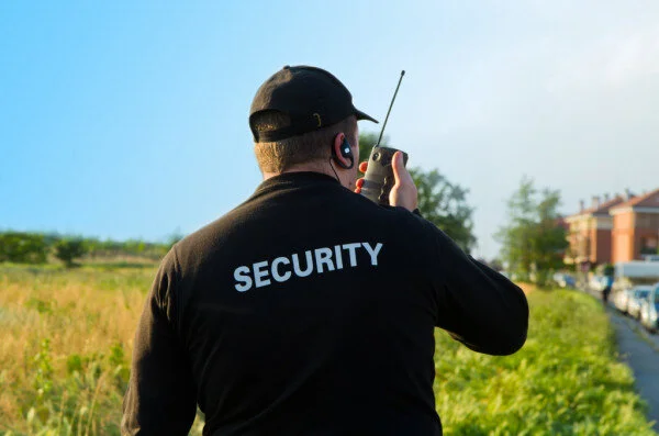 Security guard wearing a black shirt with "SECURITY" on the back, holding a walkie-talkie outdoors during daytime.