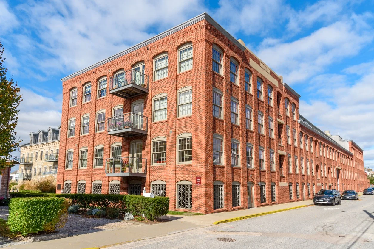 Red brick multi-story residential building with balconies, large windows, and a small front yard with bushes, located on a city street under a partly cloudy sky.