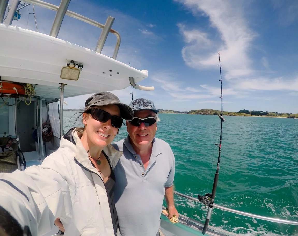 Two people taking a selfie on a boat with fishing gear in the background, ocean, and islands under a partly cloudy sky.