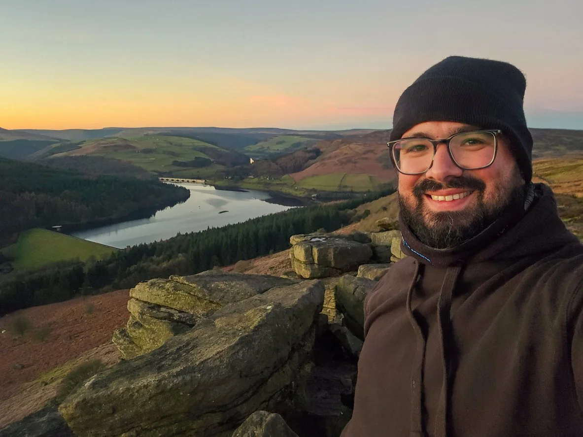 A smiling man wearing glasses, a black beanie, and a brown jacket taking a selfie on a rocky hilltop during sunset with a view of a river, green hills, and a sky with pink and orange hues.