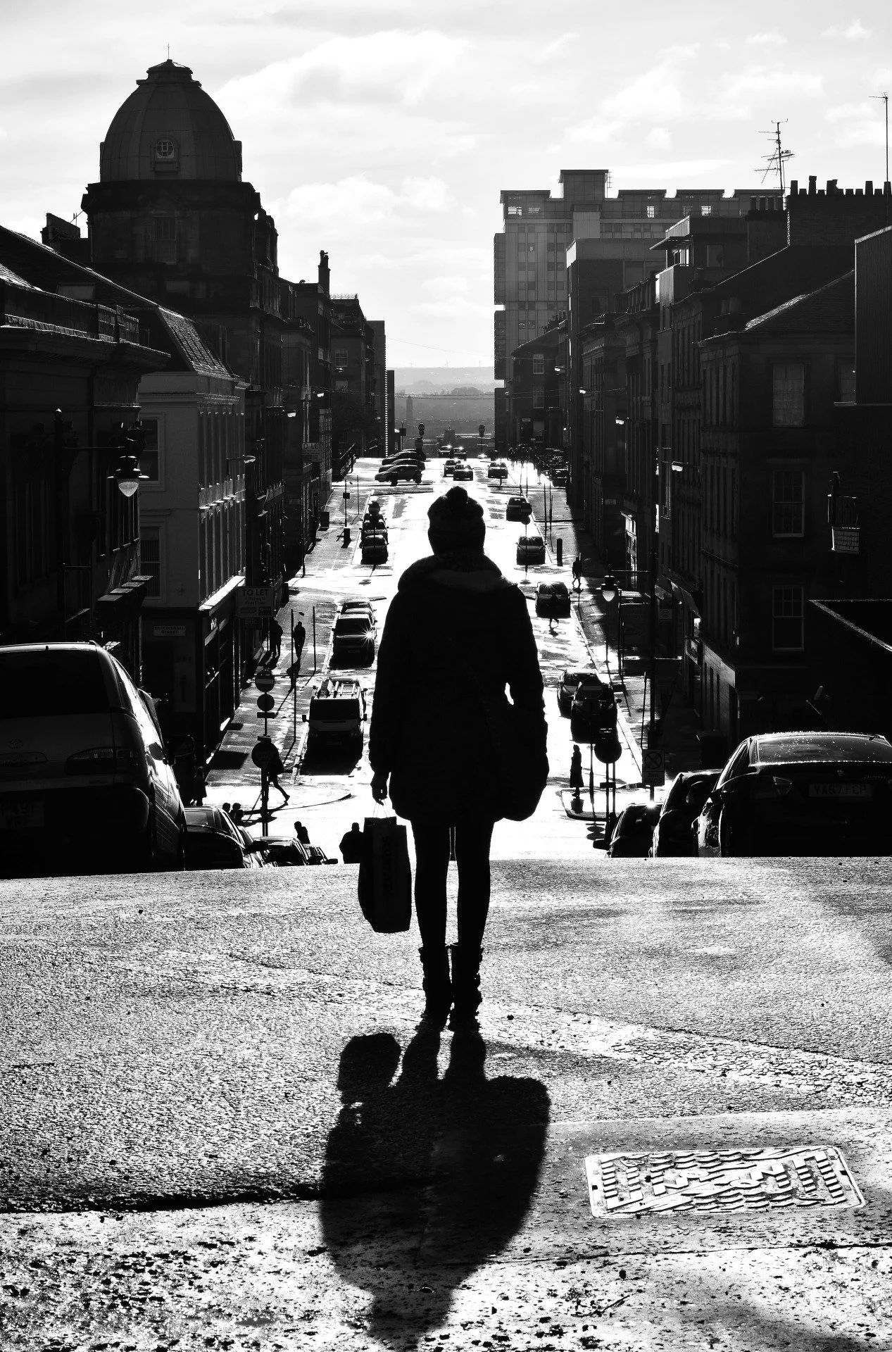 A silhouette of a woman with a bag and suitcase walking down a city street with cars parked along the sides, with buildings and a domed structure in the background, in black and white.