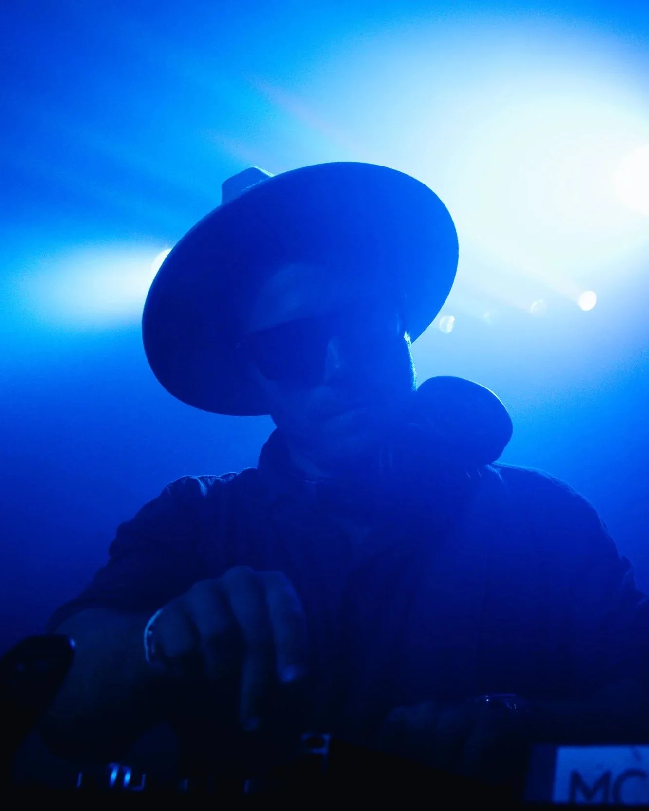 A person wearing a wide-brimmed hat and sunglasses, silhouetted against bright blue stage lights, possibly at a concert or nightclub.
