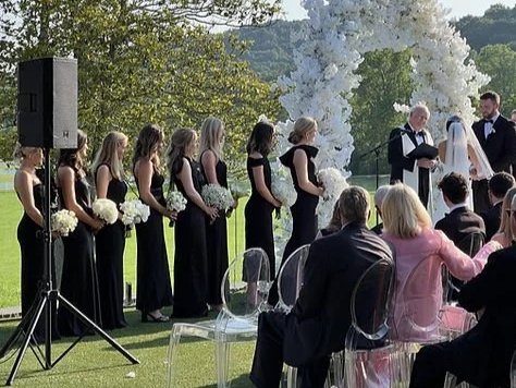 A wedding ceremony outdoors with bridesmaids in black dresses holding white bouquets and the bride and groom standing under white flowering trees, guests seated nearby.