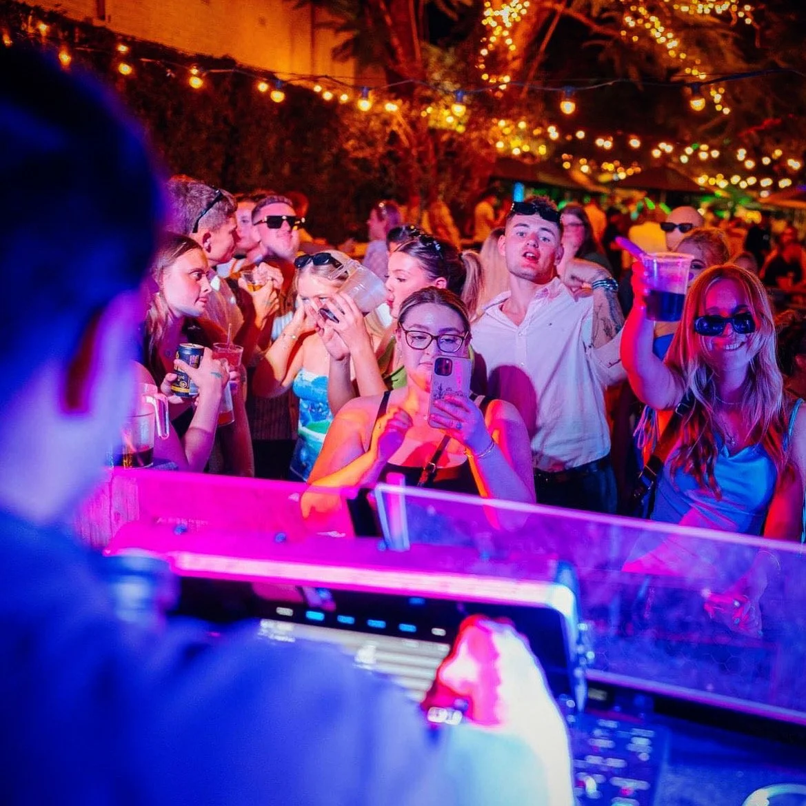 People dancing and socializing at an outdoor party with string lights and colorful lighting, some wearing sunglasses and holding drinks.