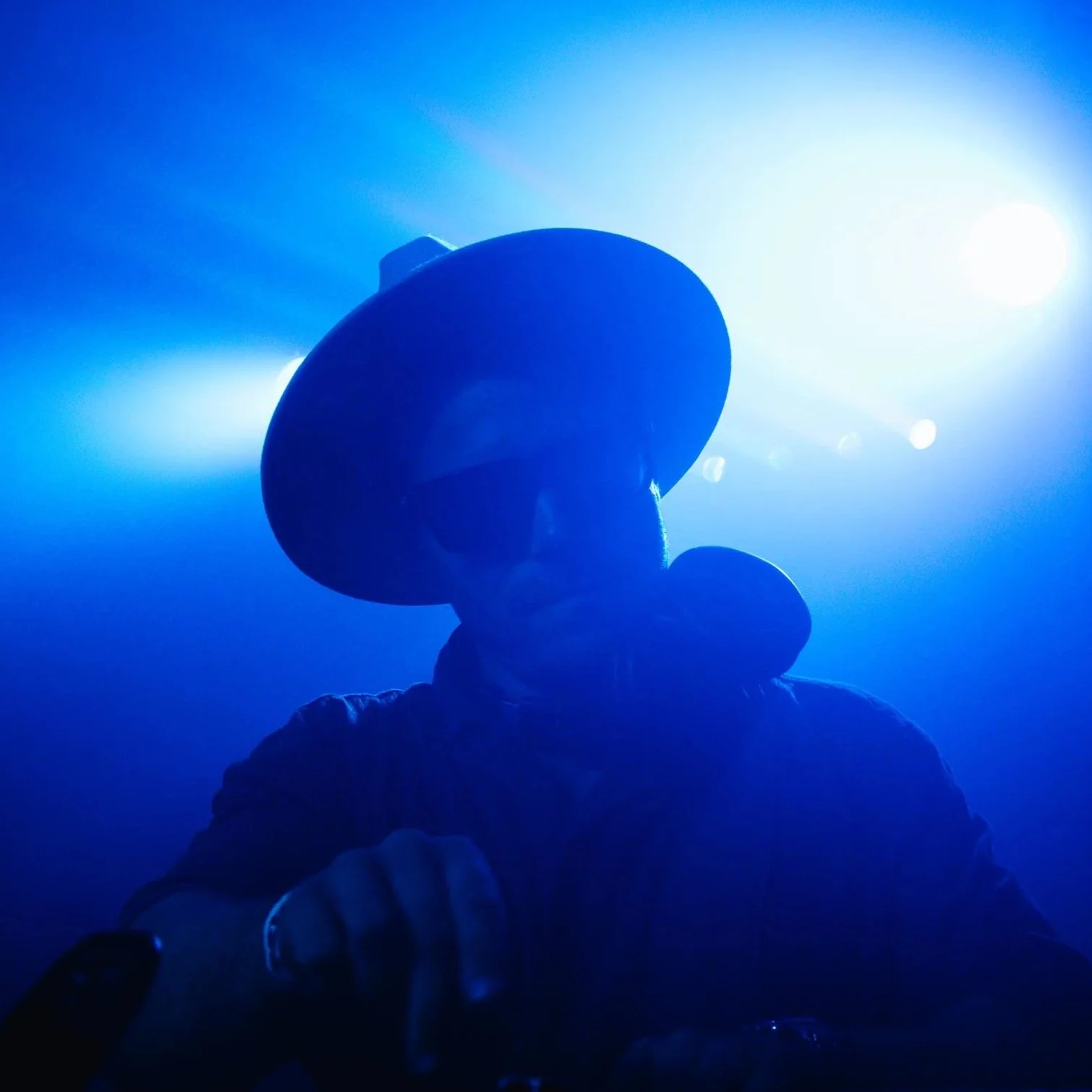 Person wearing a wide-brimmed hat and sunglasses, backlit by bright blue lights, creating a silhouette effect.
