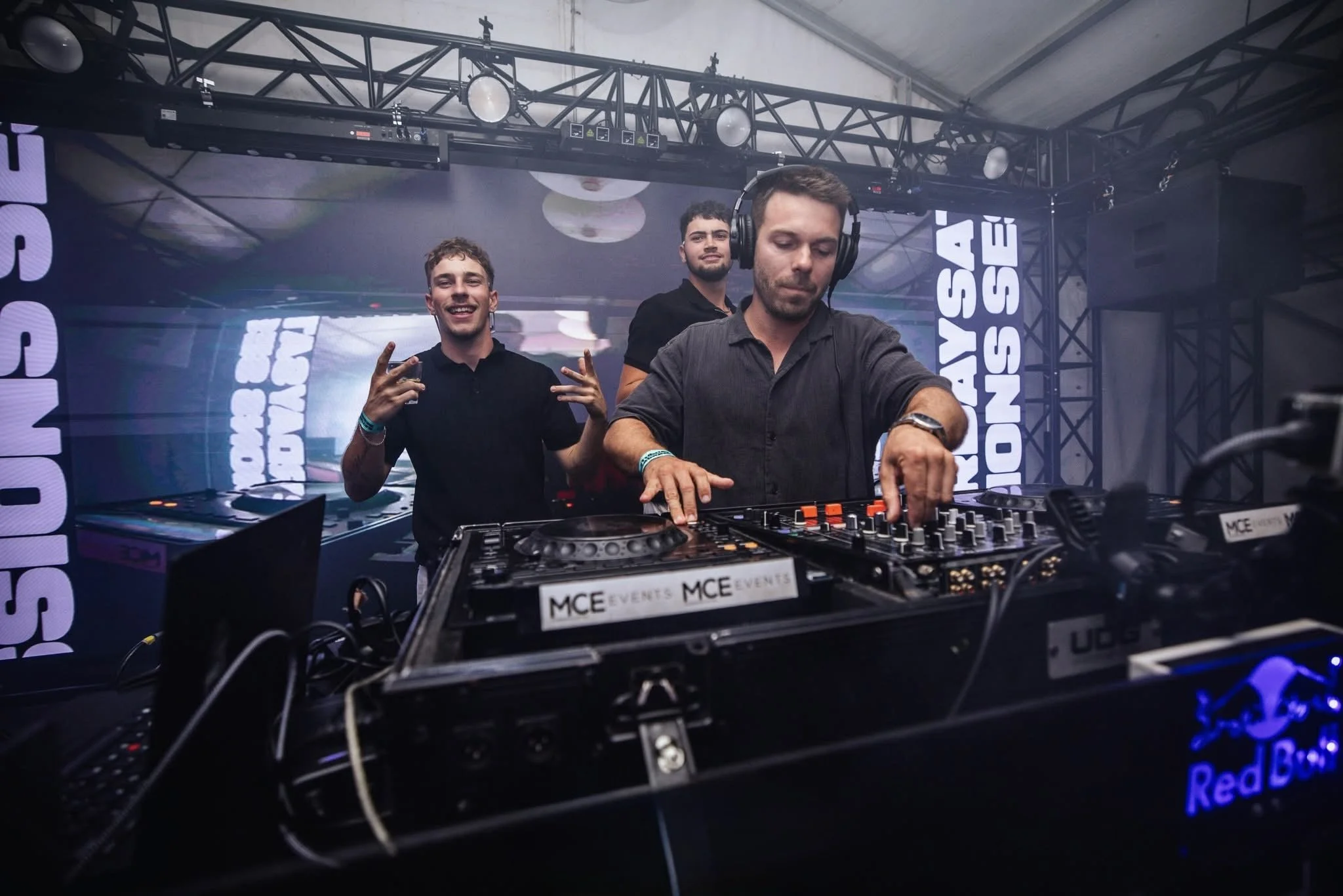 Three men at a DJ booth, one wearing headphones and operating the DJ equipment, and the other two posing and smiling at the camera, with a stage and LED screens in the background.
