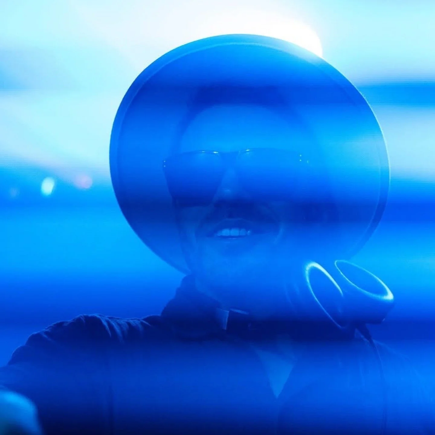 Person wearing a large hat and sunglasses, illuminated with blue lights, taking a selfie.