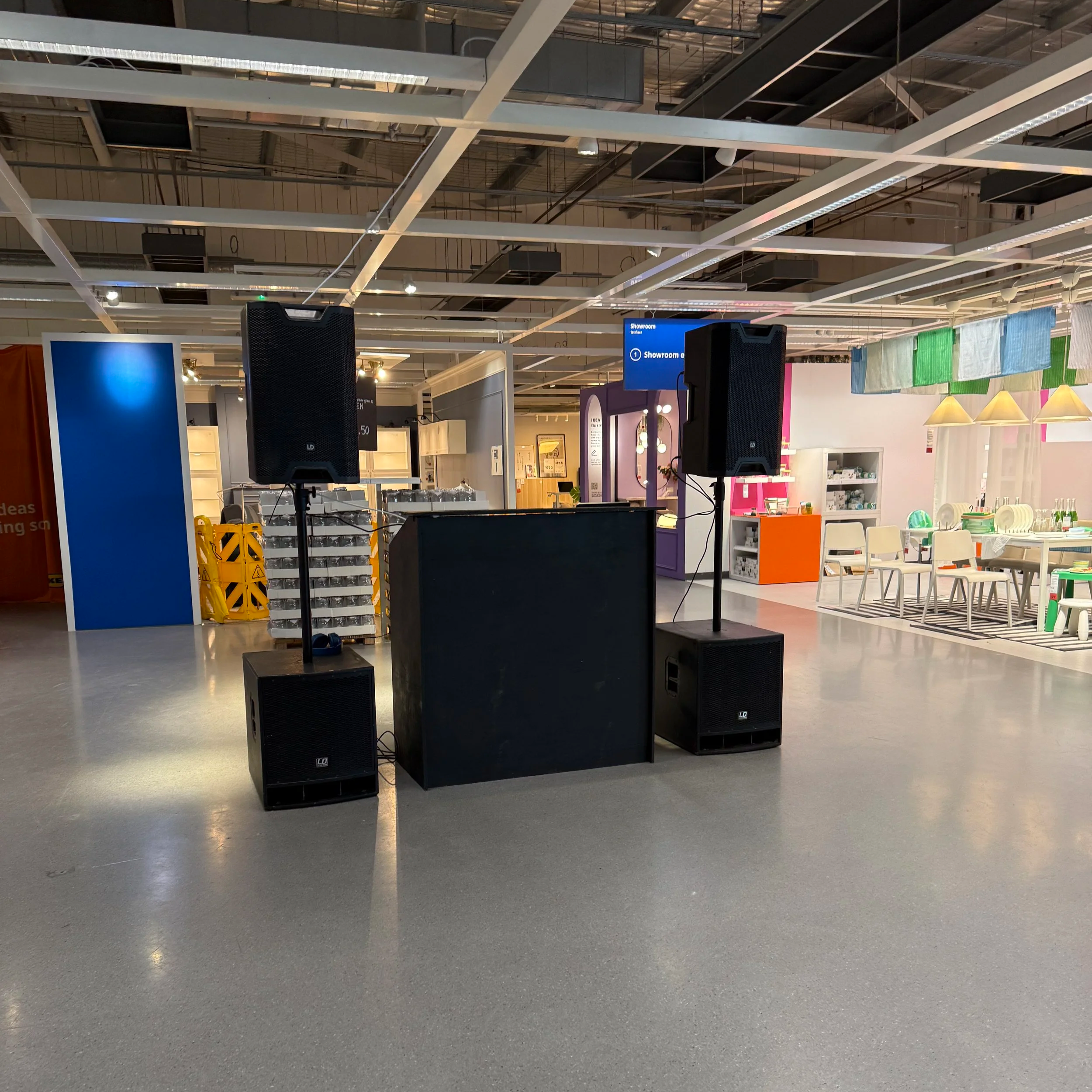 Empty indoor space with two large black speakers on stands, a DJ booth or equipment table in the center, and a colorful children's furniture display in the background.