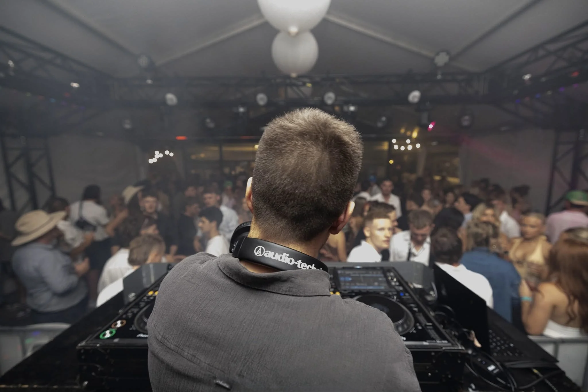 DJ performing at a crowded nightclub with people dancing and socializing under ceiling lights and fog.