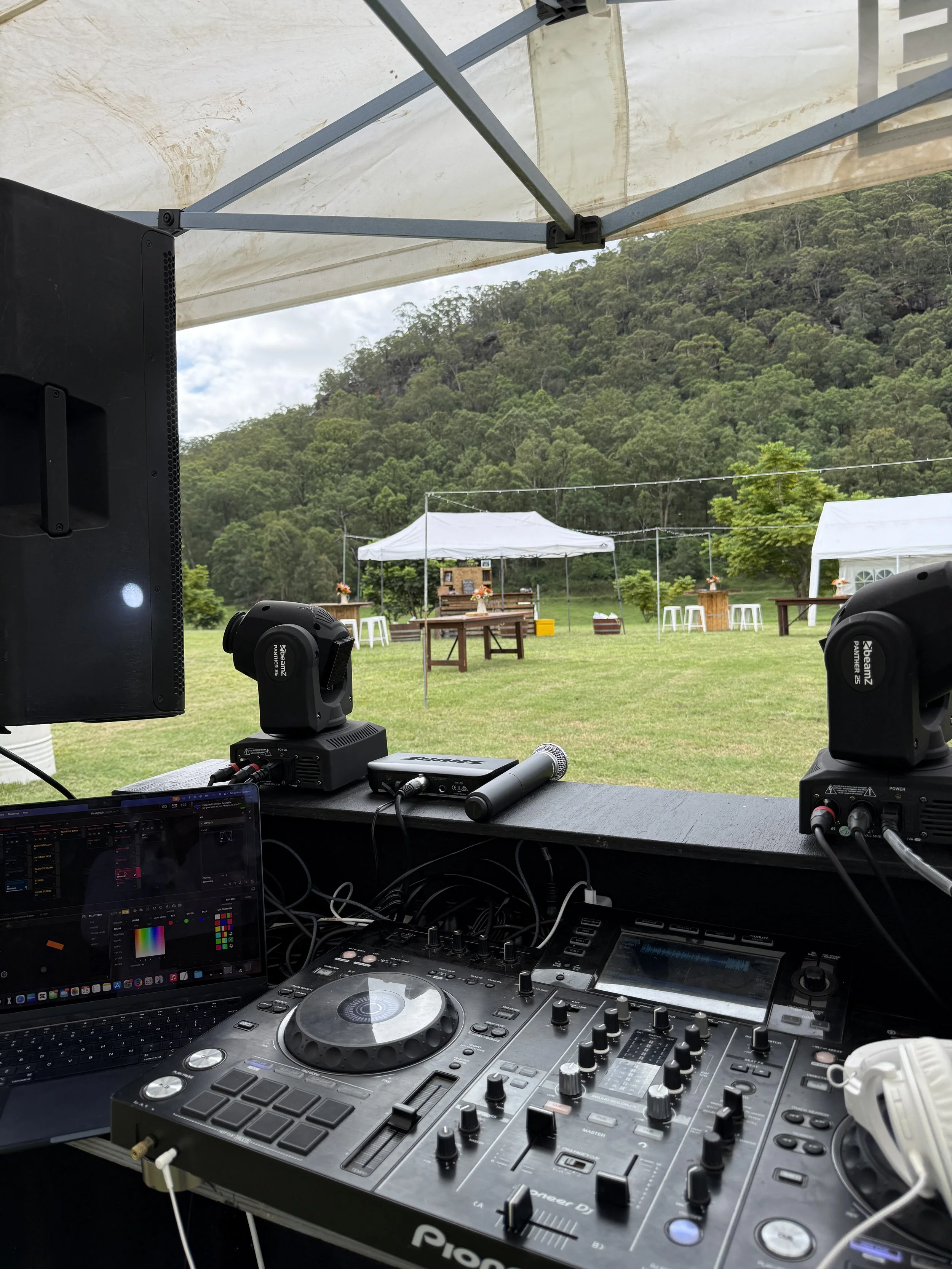 DJ equipment with turntable, mixer, laptop, and speakers set up inside a tent at an outdoor event, with a view of green hills and outdoor tables in the background.