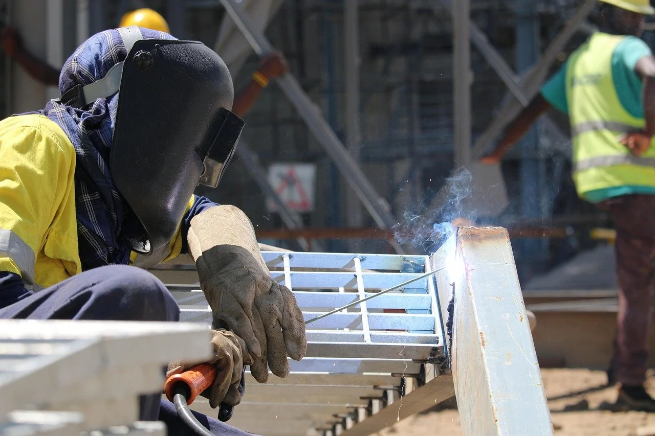 Un trabajador sombrea usando un casco y guantes mientras realiza soldadura en una estructura metálica en un sitio de construcción.