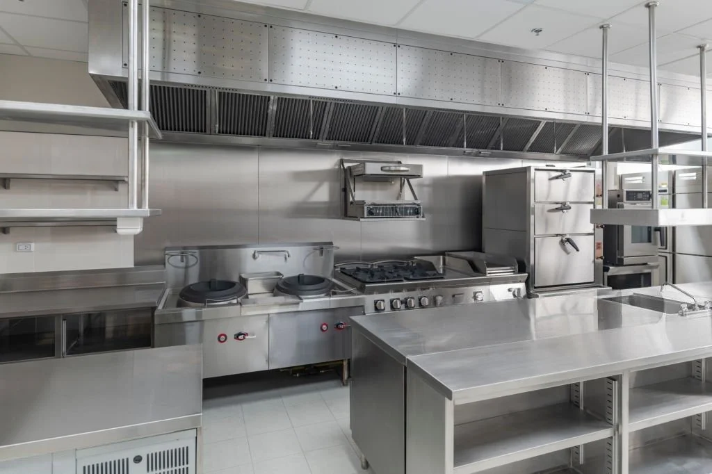Cocina profesional limpia con estanterías, estufa, horno y campana extractora de acero inoxidable.
