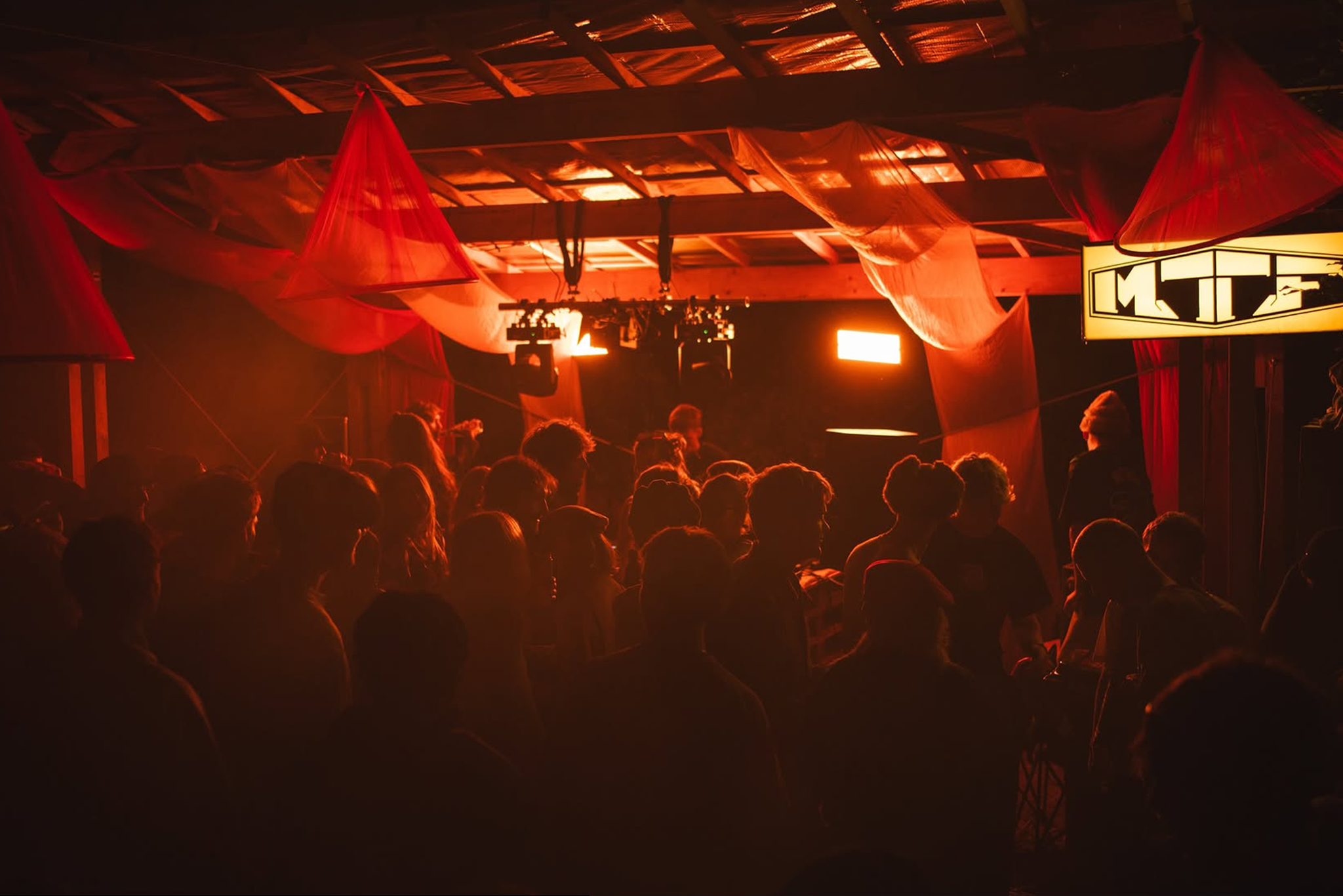 An indoor concert or event venue with red and orange lighting illuminating the ceiling drapes and audience members seated in front of a stage with spotlights and equipment visible.