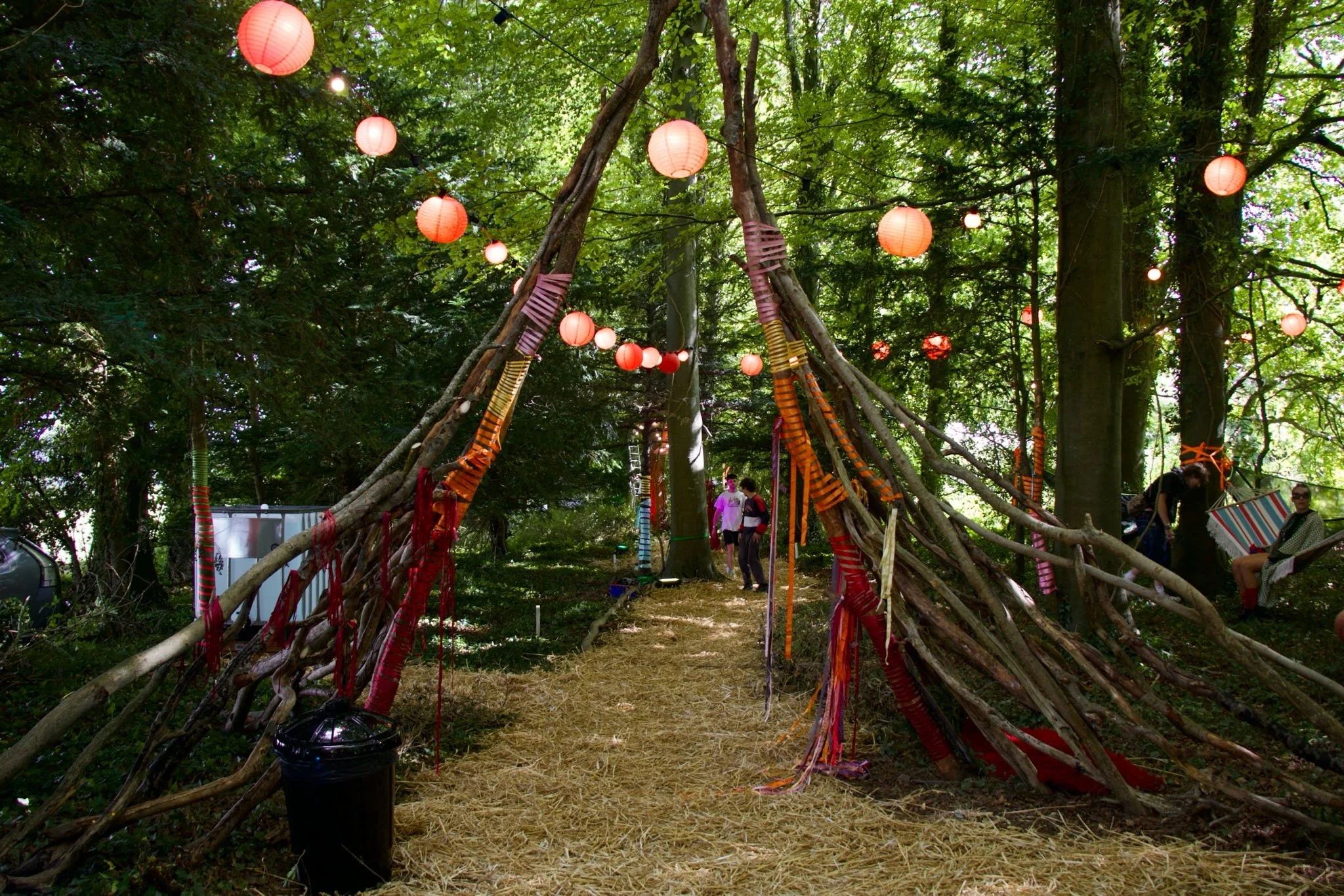 A teepee-like structure made of tree branches and tied with colorful fabric strips, decorated with red paper lanterns hanging from string lights in a green outdoor forest setting, with people gathered nearby.
