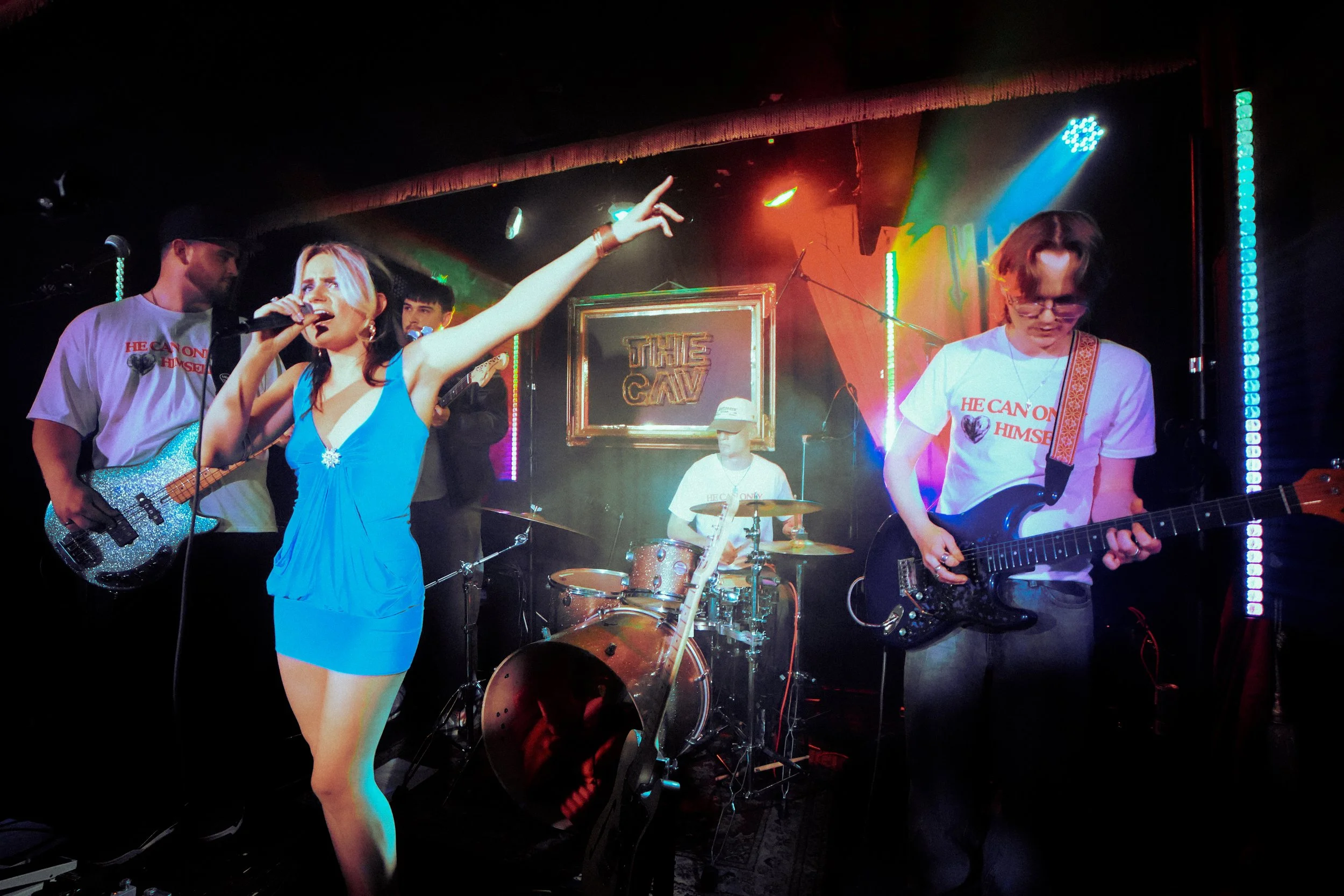 A band performing live on stage, including a female singer in a blue dress holding a glittery guitar, a guitarist on the right, a drummer in the background, and two men wearing white T-shirts with red and black text. The stage is lit with colorful lights and features a framed sign that reads 'THE CAV'.