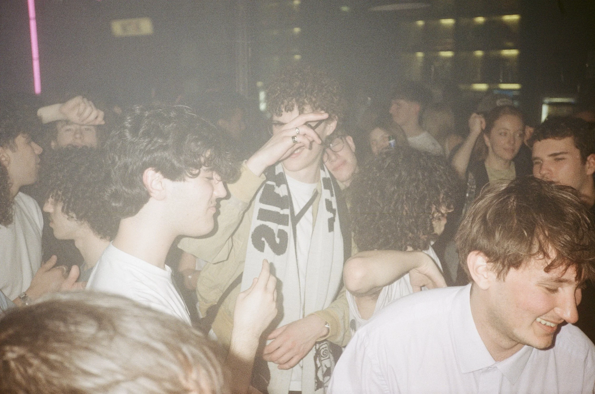 A group of young people at a party, crowded together, some smiling, one person adjusting or touching their glasses, with a lively atmosphere and multiple people in the background.